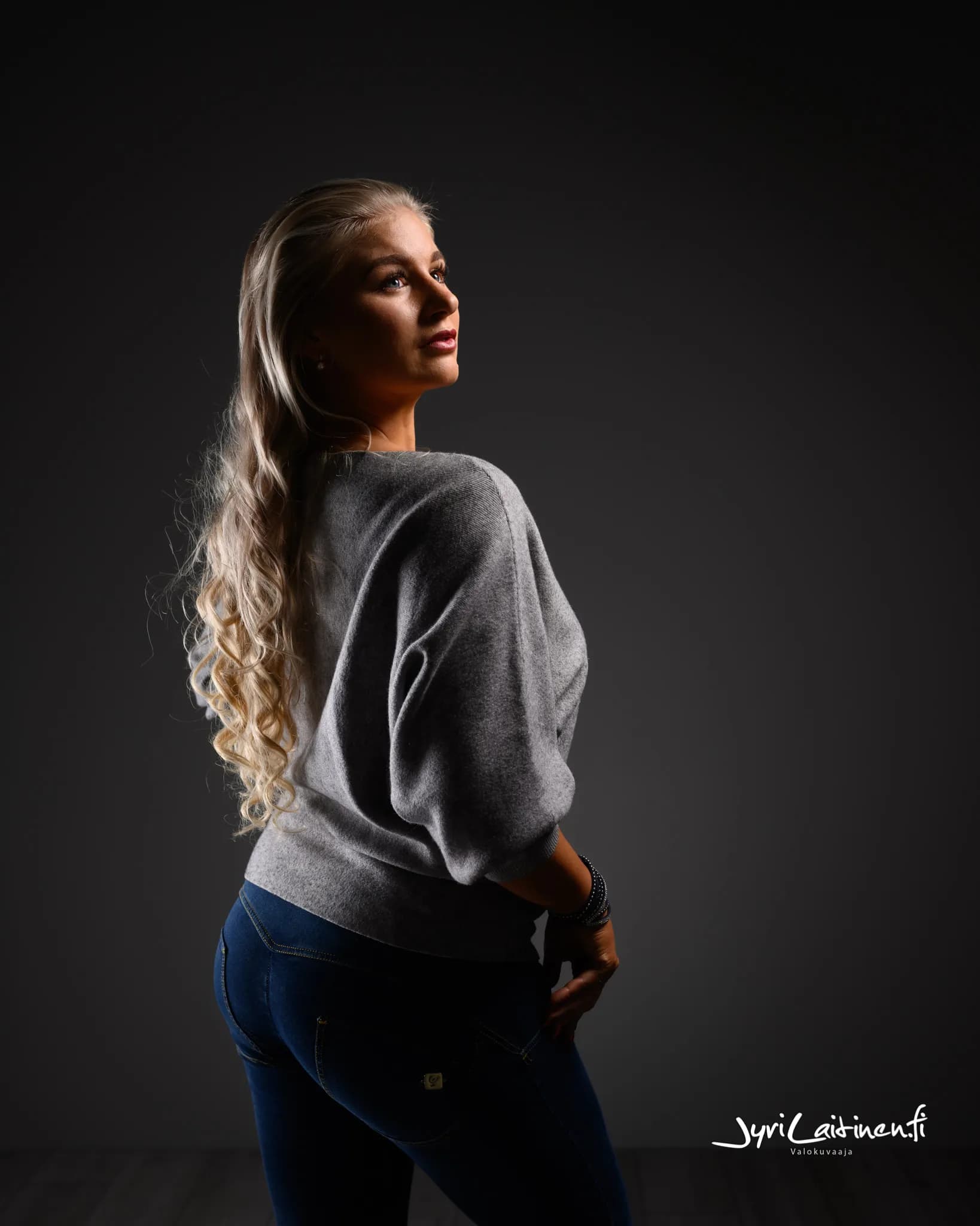 Emily posing in a gray sweater and jeans against a dark background.