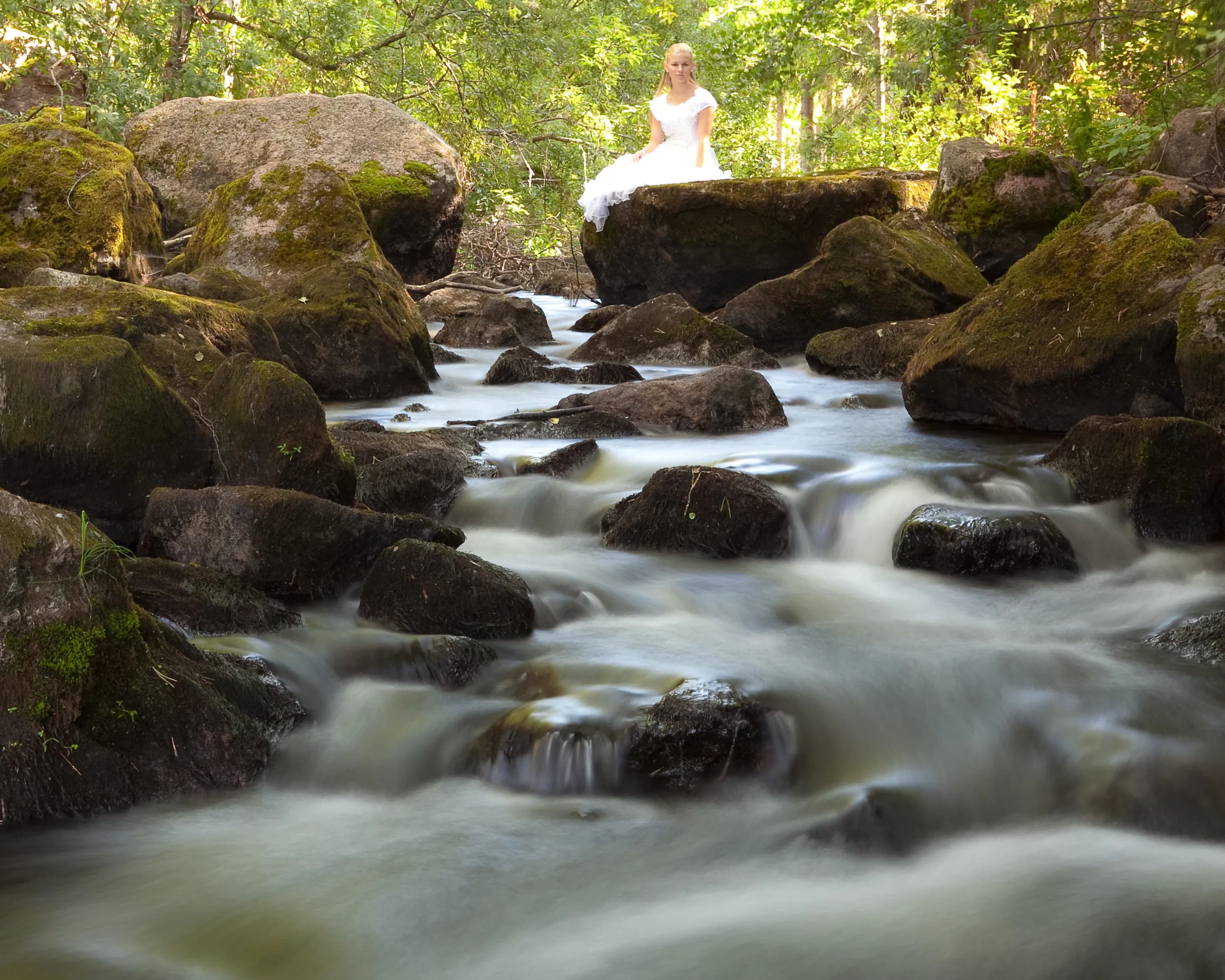 Emily in a white dress sitting on a rock by a flowing stream in a forest.