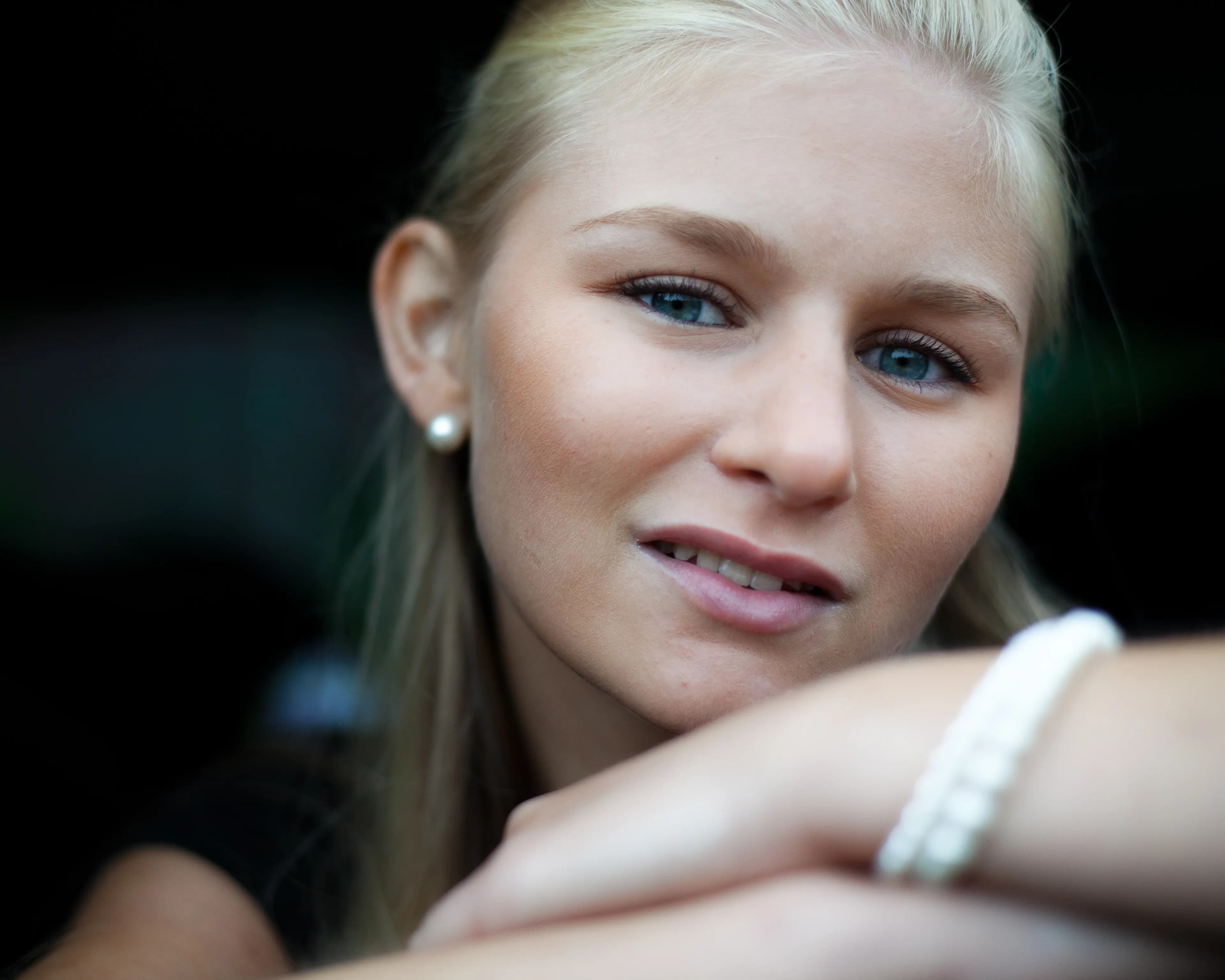 Emily with long blonde hair, wearing pearl earrings and a bracelet, looking at the camera.