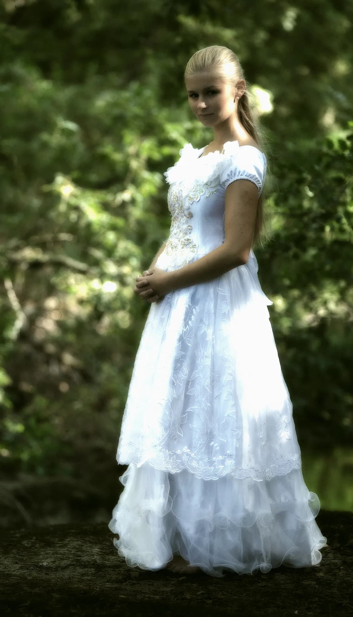 Emily in a white, elegant dress standing outdoors with greenery in the background.