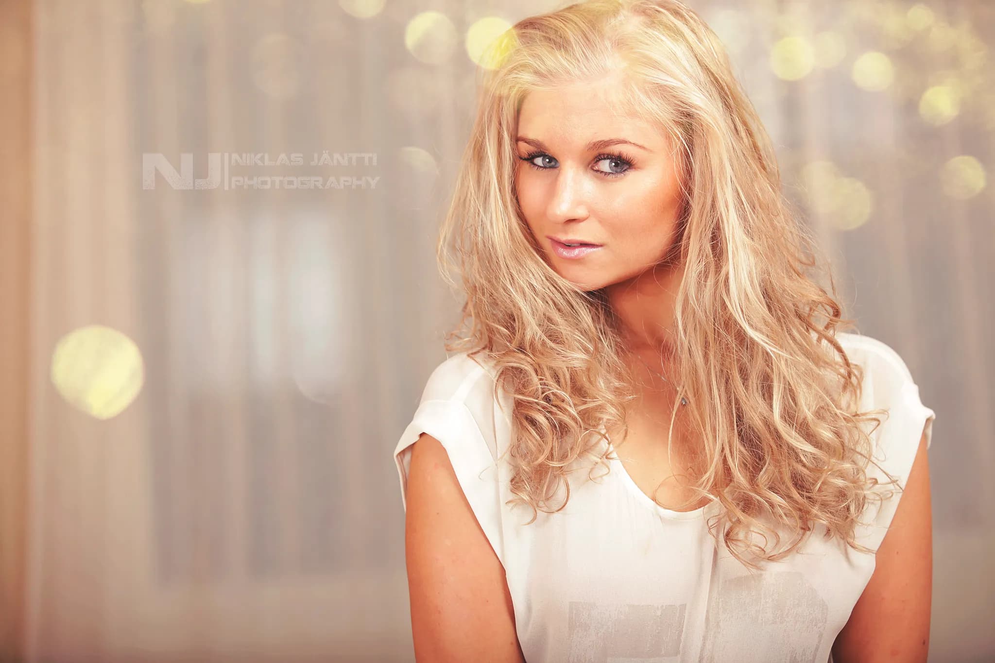 Emily with long, wavy blonde hair, wearing a white top, posing against a softly lit background.