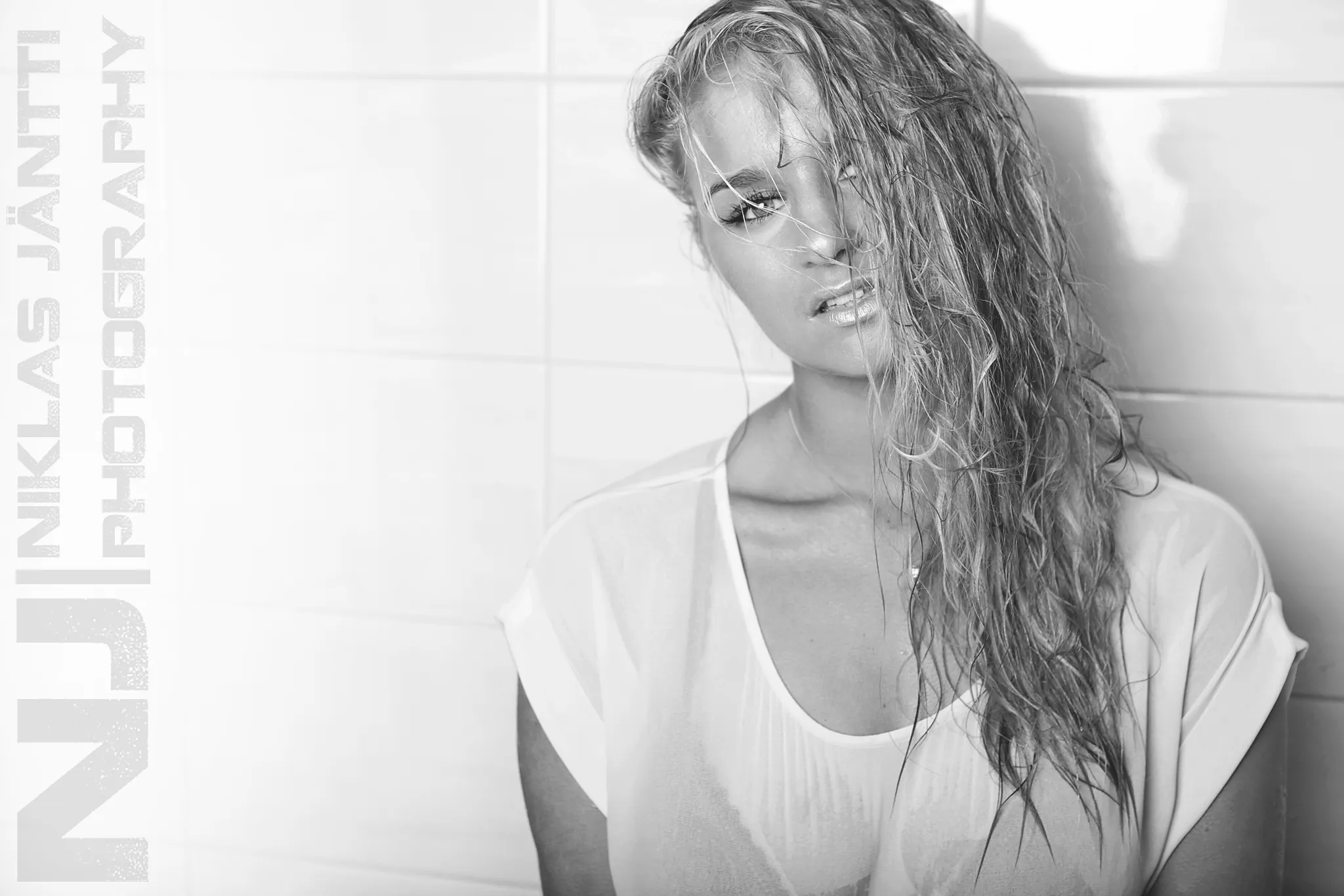 Emily posing with wet hair against a tiled wall, wearing a sheer top.