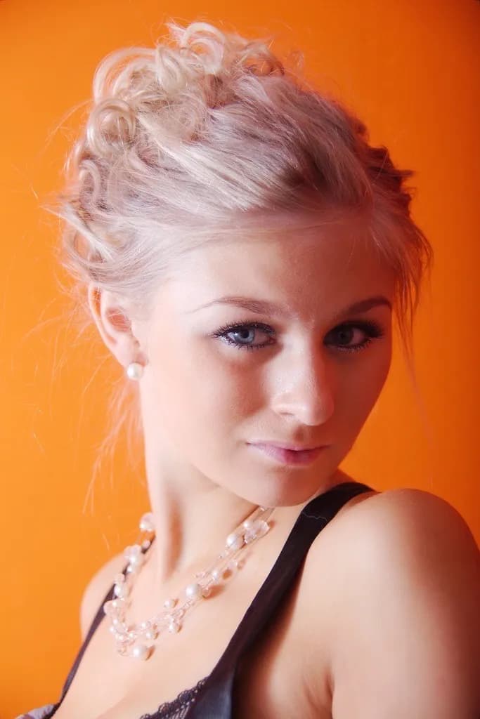 Emily with blonde hair styled in an updo, wearing a pearl necklace against an orange background.