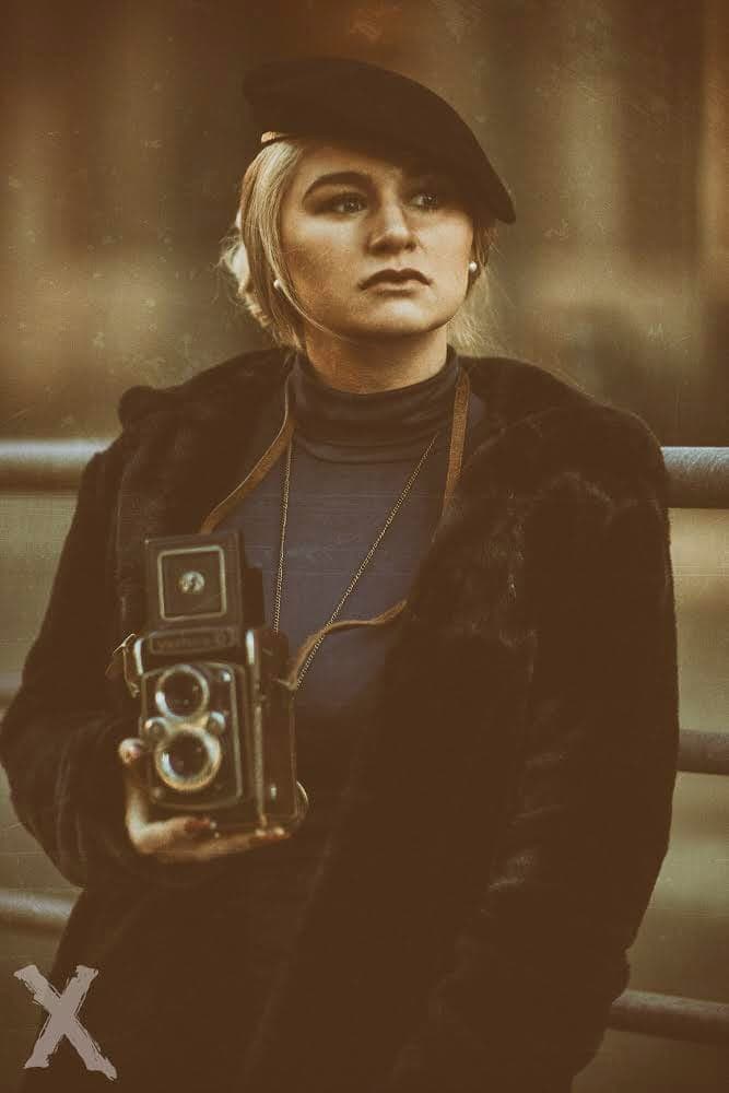 Emily holding a vintage camera, wearing a beret and a dark coat.