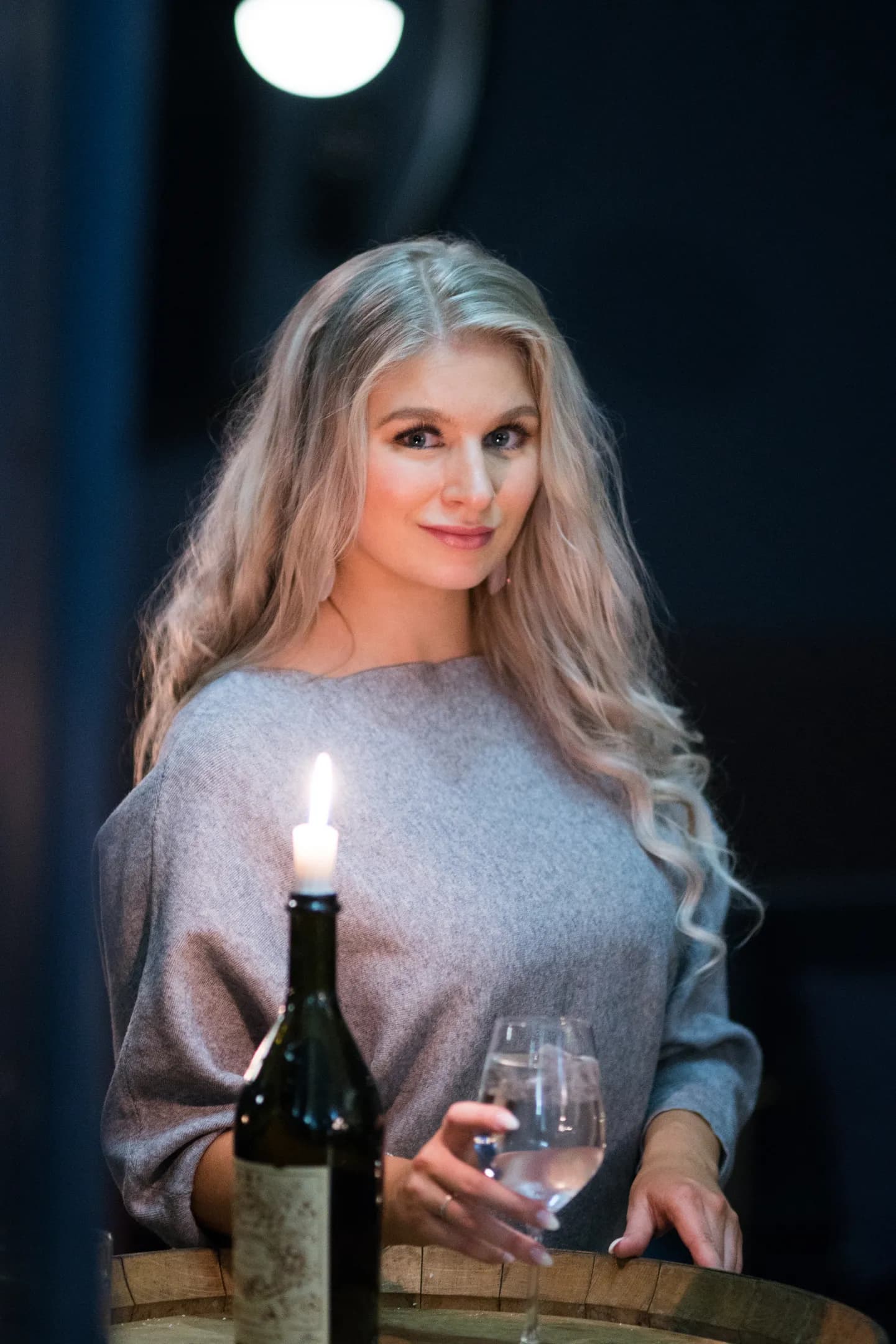 Emily with long blonde hair, wearing a gray top, holding a wine glass next to a lit candle.