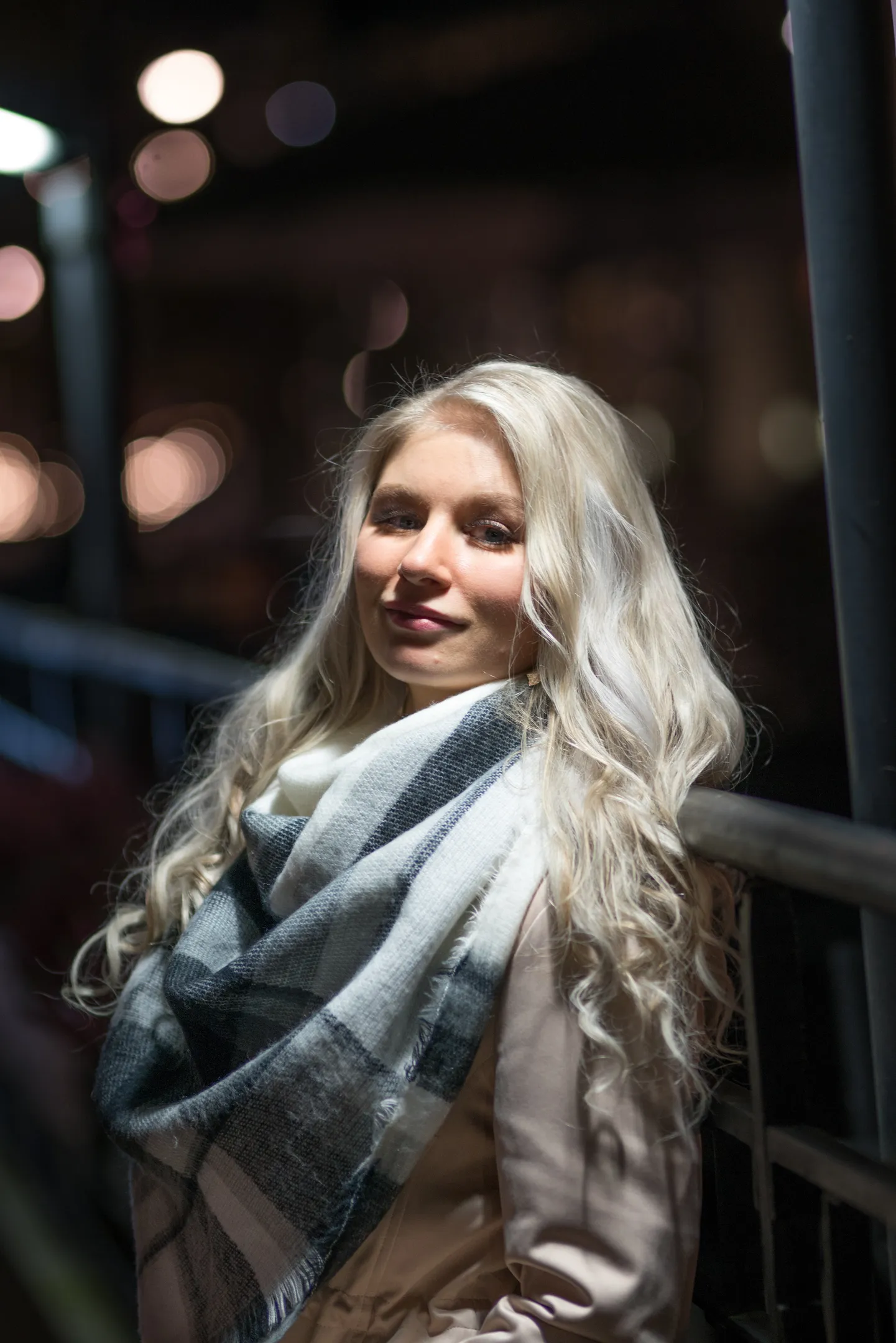 Emily with long blonde hair, wearing a plaid scarf, standing by a railing at night.