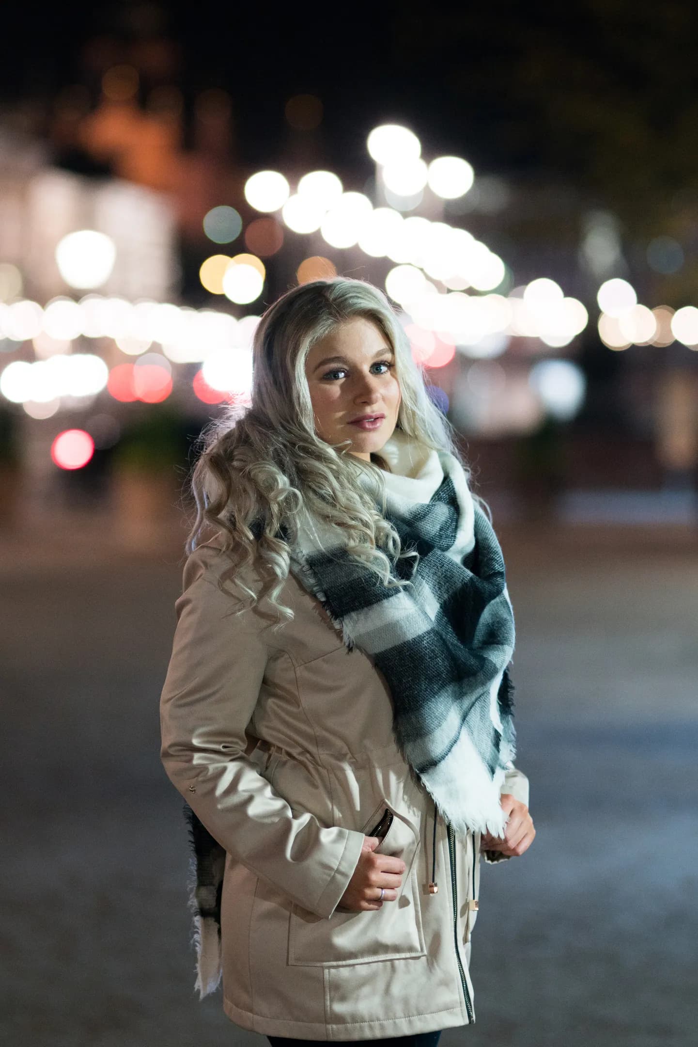 Emily standing outdoors at night, wearing a beige coat and a plaid scarf.