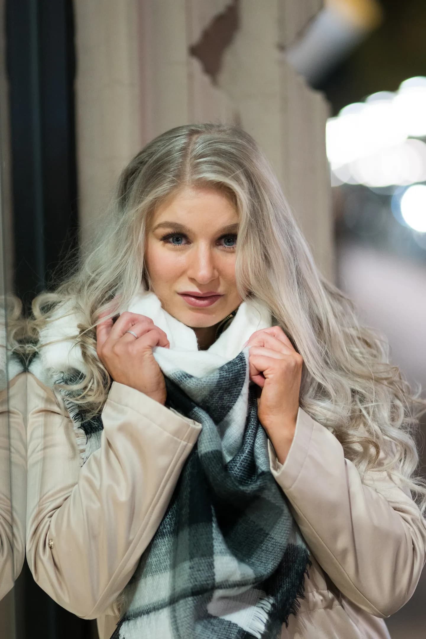 Emily with long blonde hair, wearing a beige coat and a plaid scarf, standing outdoors at night.