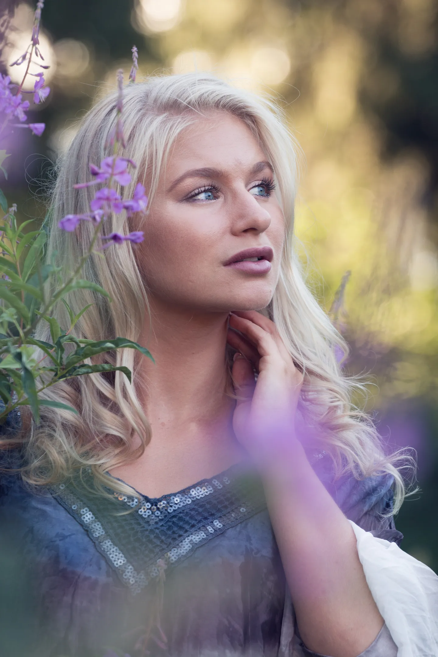 Emily with long blonde hair, surrounded by purple flowers, looking thoughtfully to the side.
