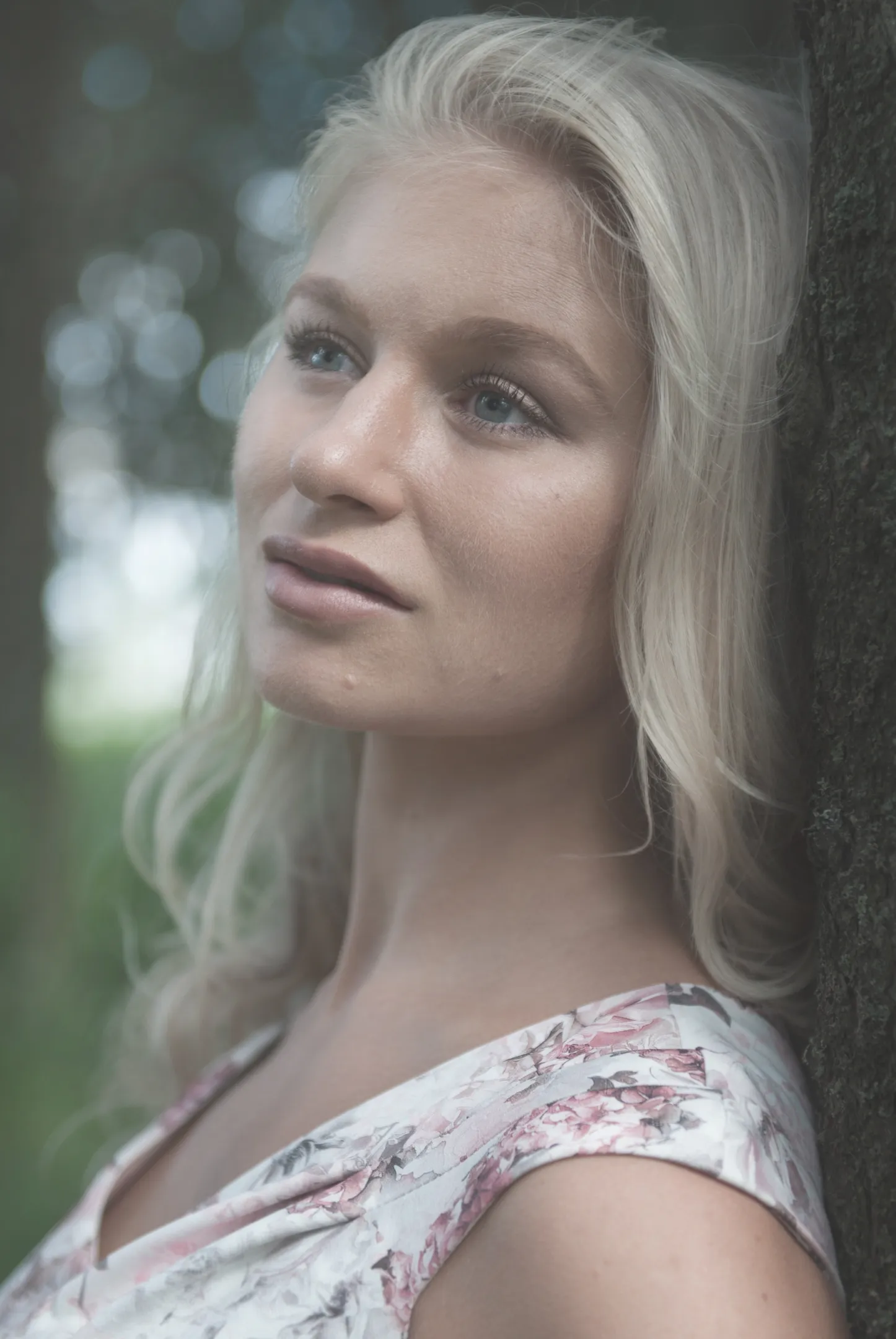 Emily with long blonde hair, wearing a floral dress, leaning against a tree in a serene outdoor setting.