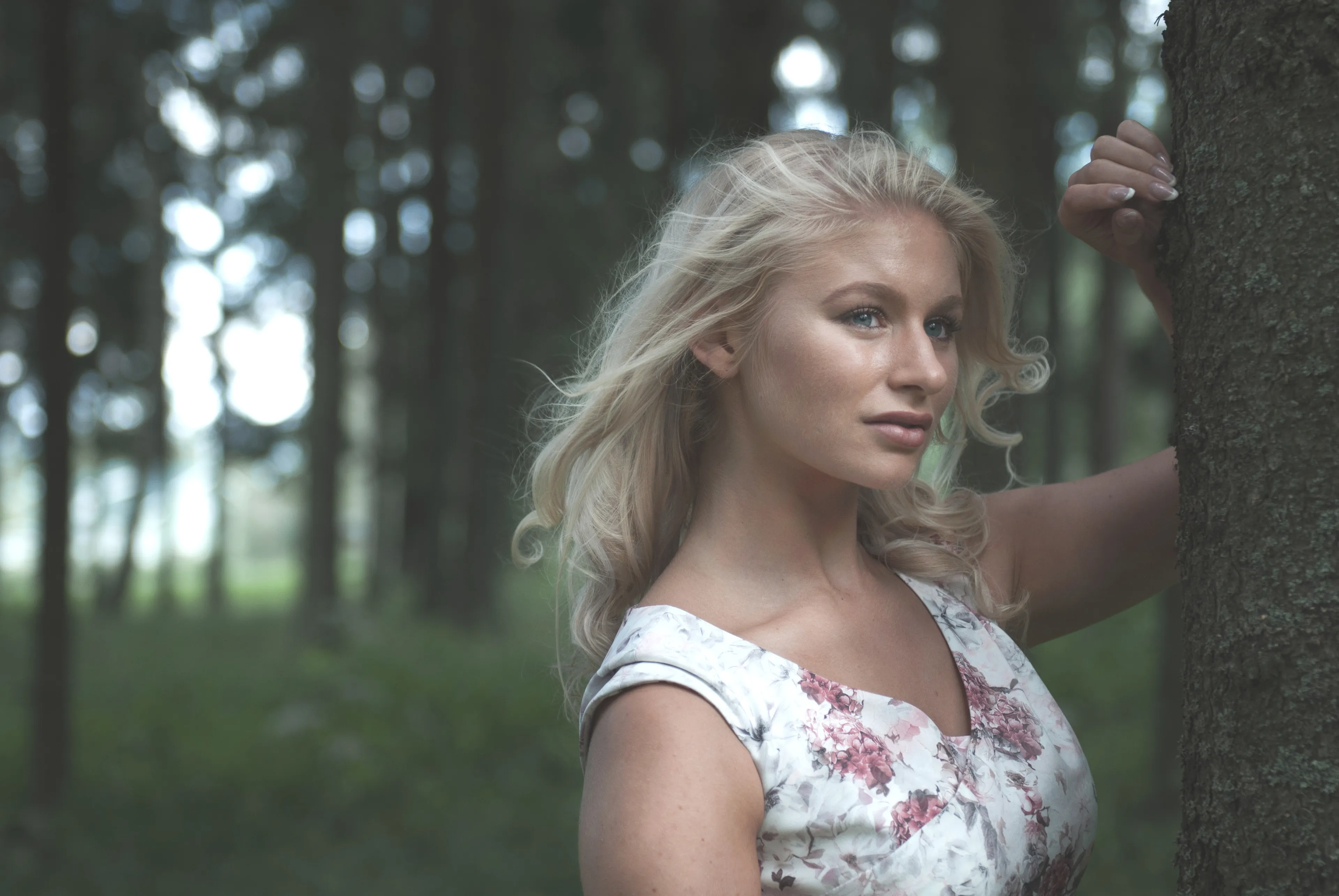 Emily standing by a tree in a forest, wearing a floral dress.