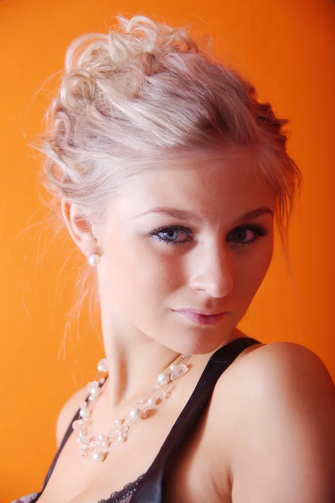 Emily with blonde hair styled in an updo, wearing a pearl necklace against an orange background.