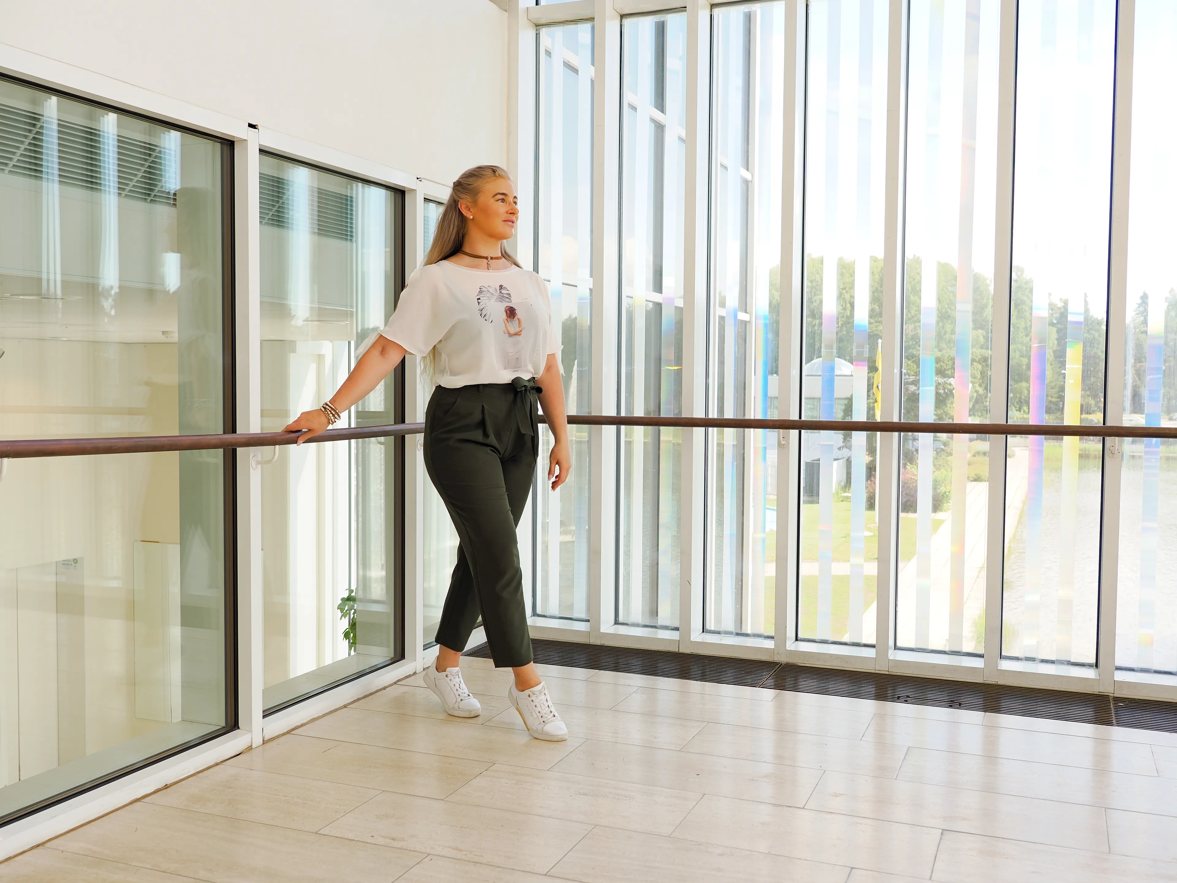 Emily standing in a modern building, wearing a white shirt and dark pants.