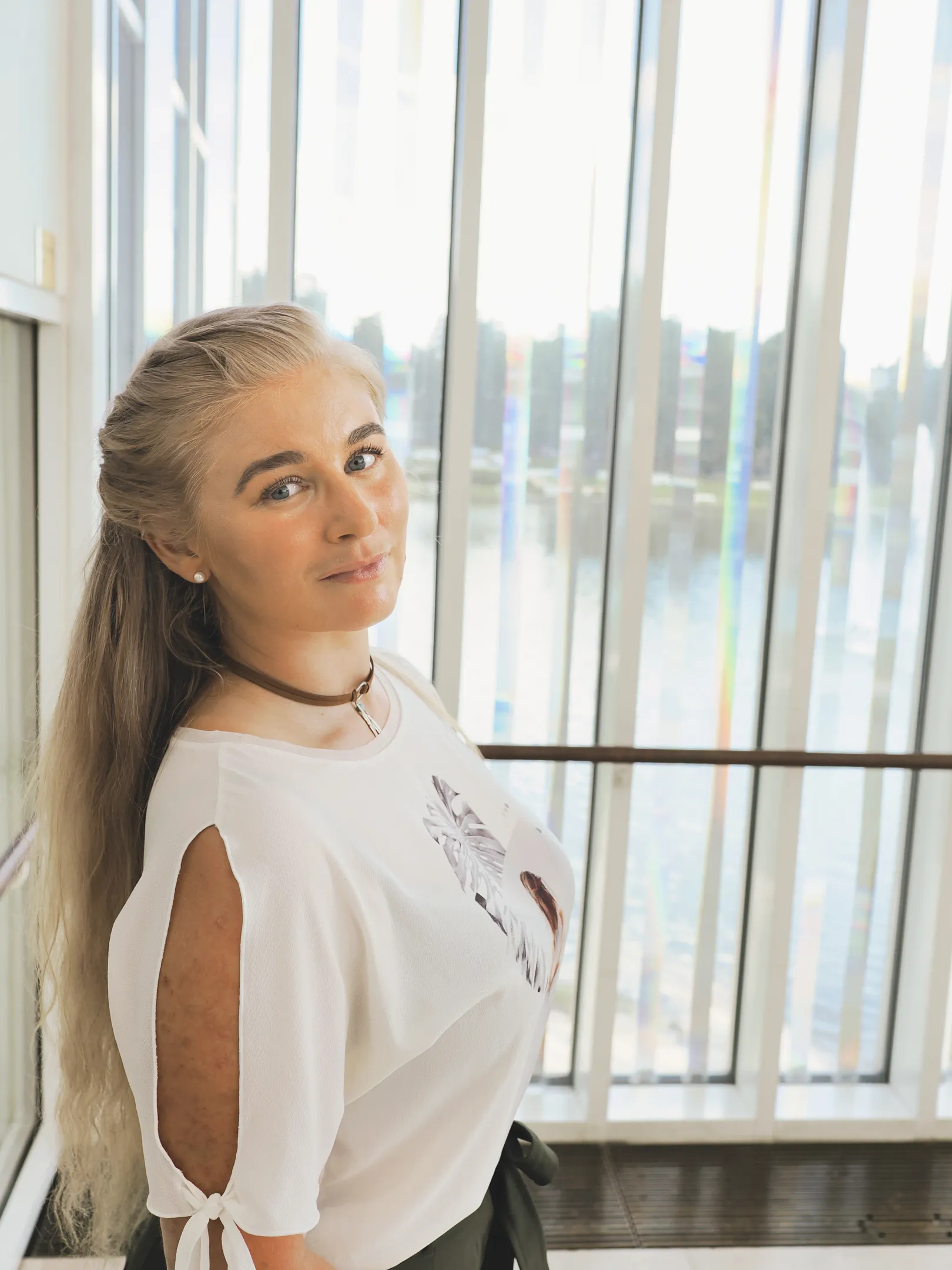 Emily standing by a window with long hair and a white top.