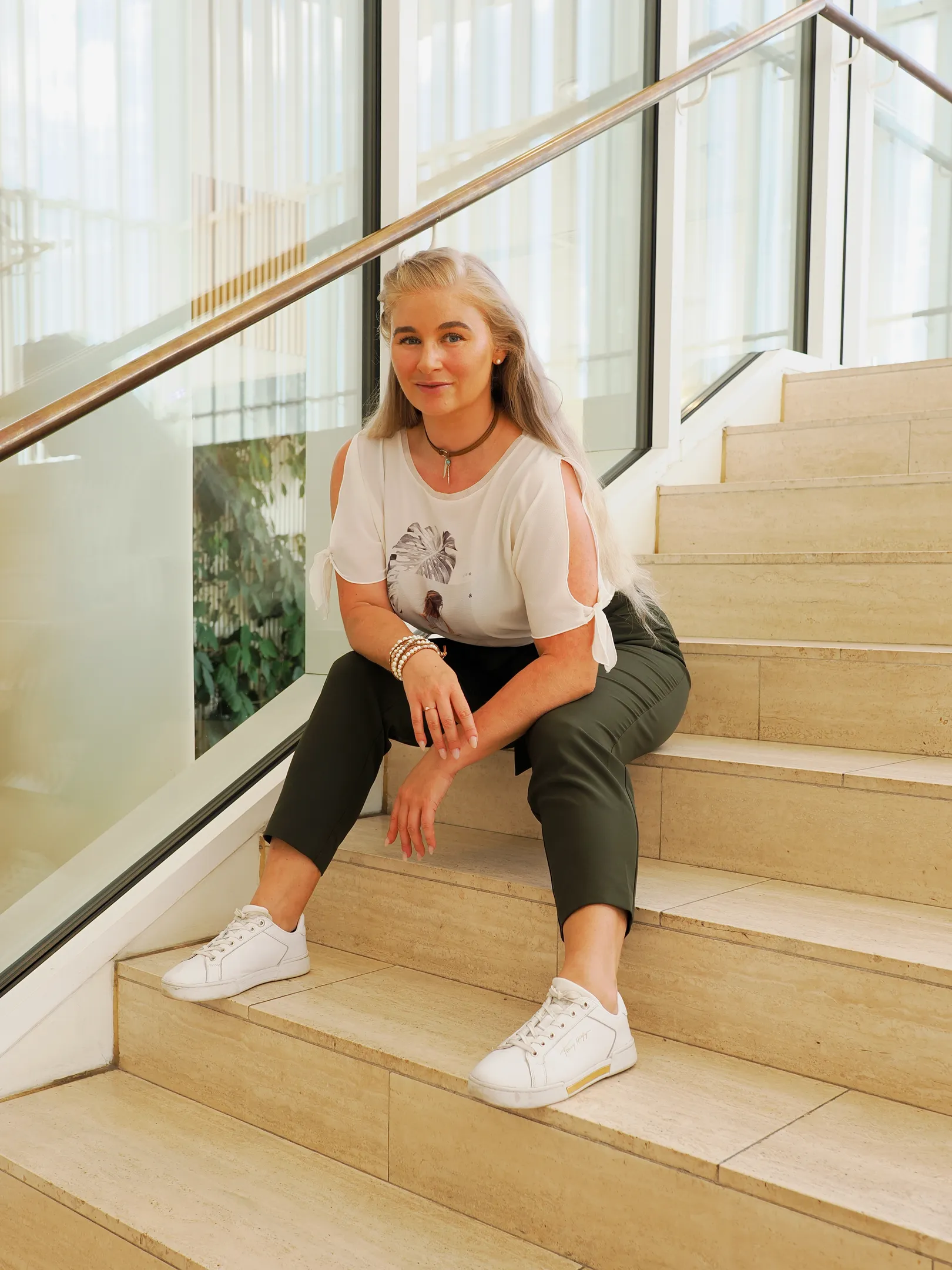 Emily sitting on stairs in a casual outfit with a white top and green pants.