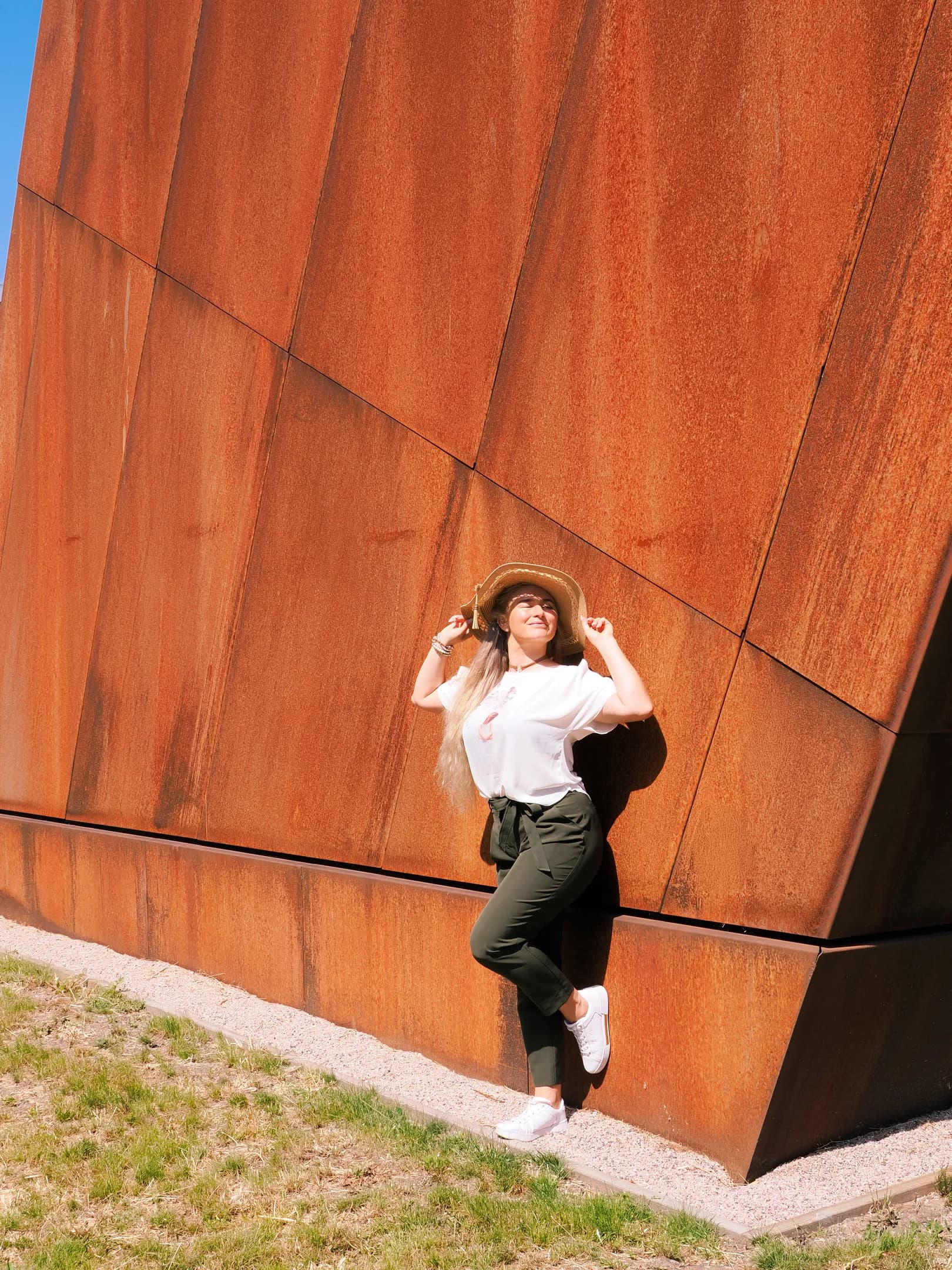 Emily posing against a large rust-colored metal wall, wearing a white shirt, green pants, and a hat.