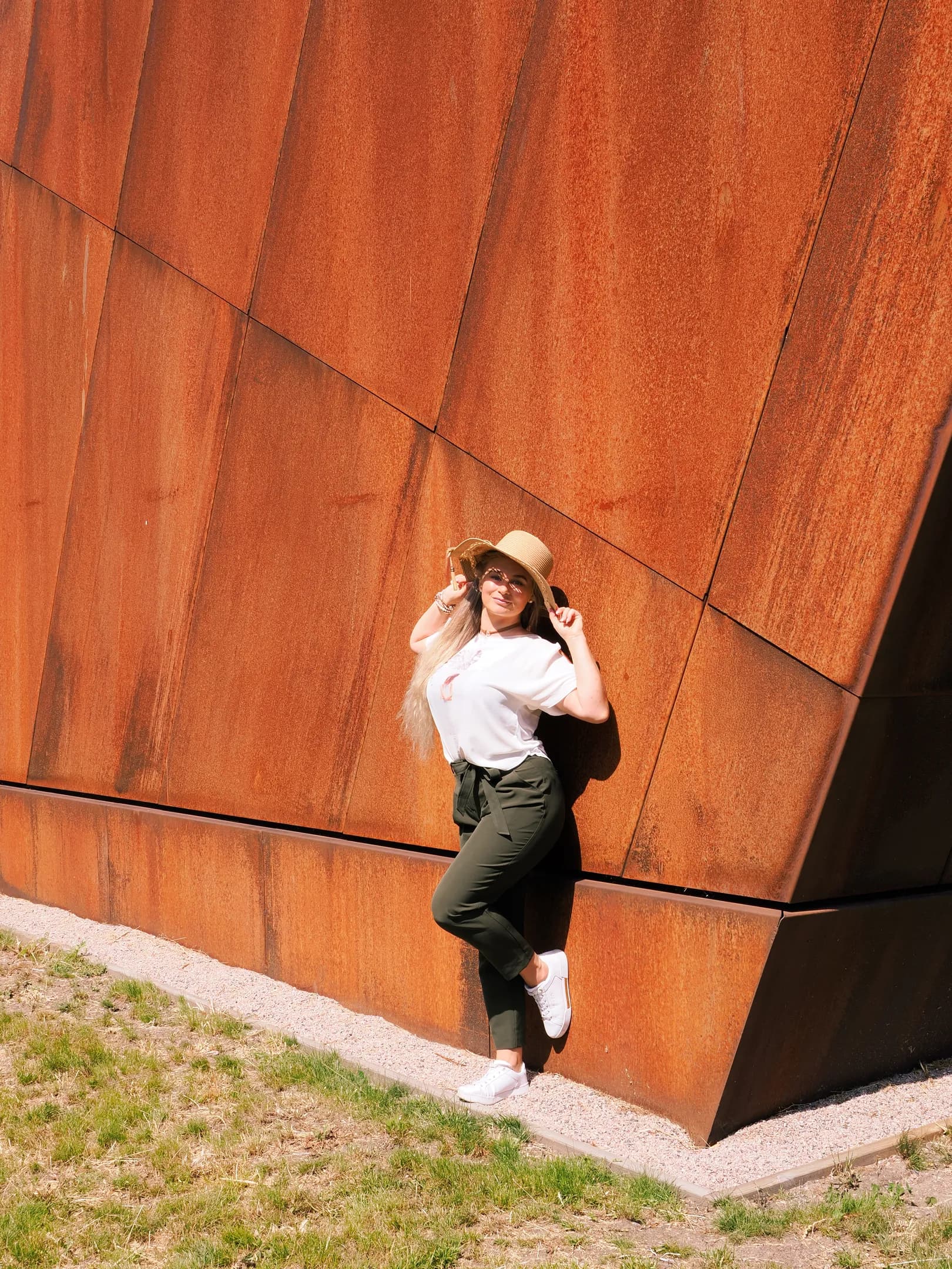 Emily posing against a rust-colored geometric wall, wearing a white top, green pants, and a hat.