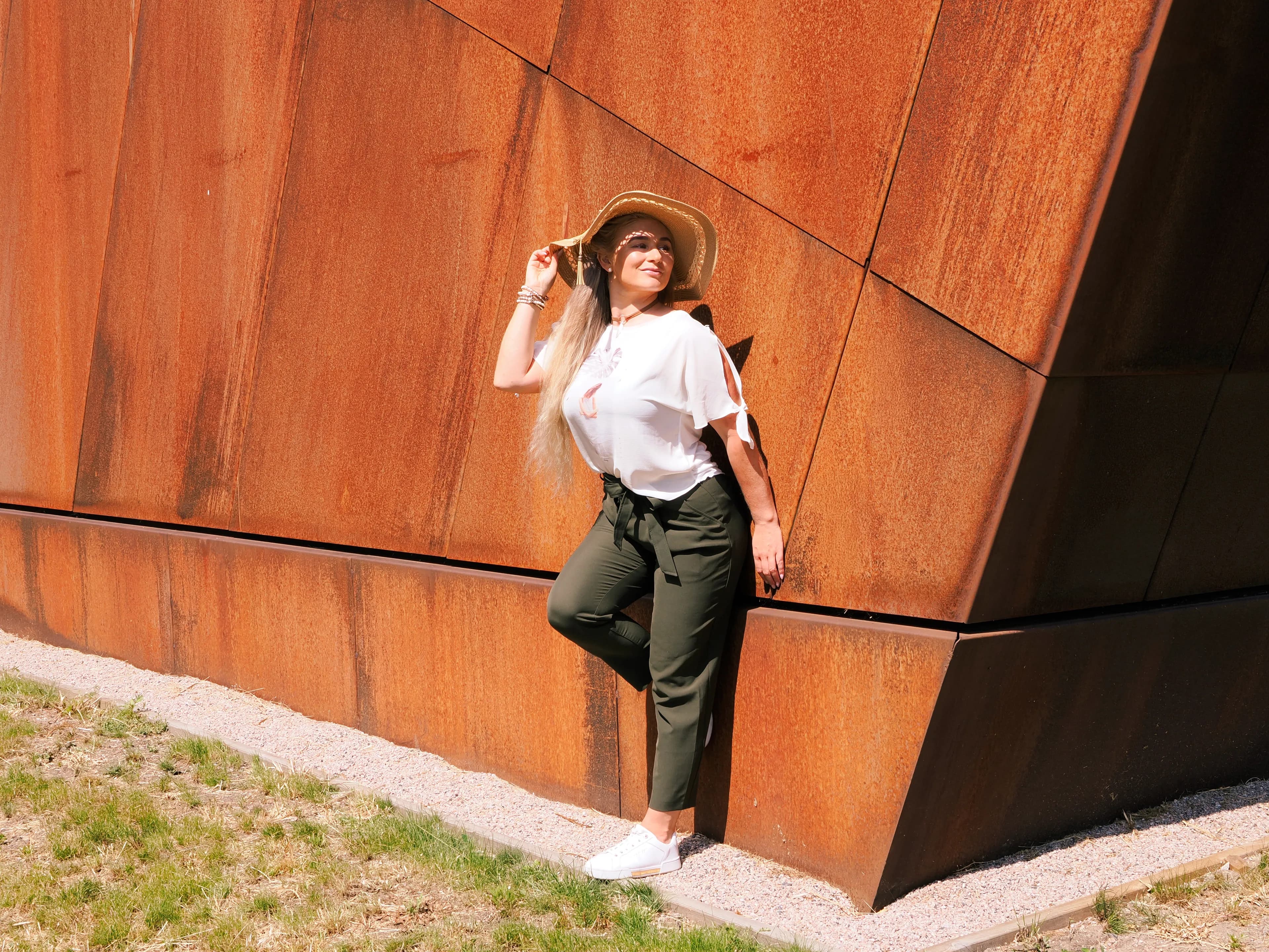 Emily posing against a rust-colored wall, wearing a white top, green pants, and a straw hat.