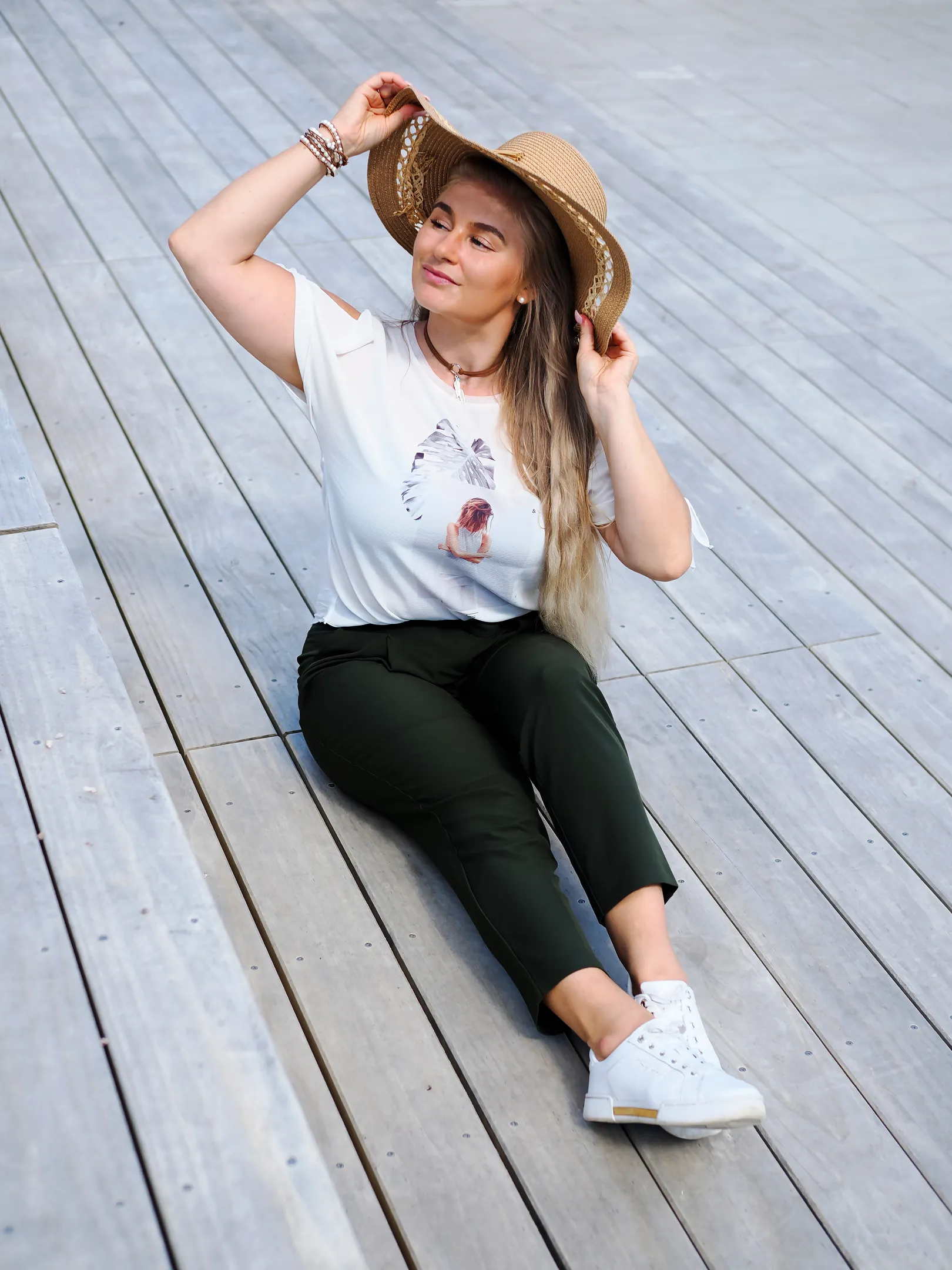 Emily sitting on wooden planks, wearing a white t-shirt, dark pants, white sneakers, and a straw hat.