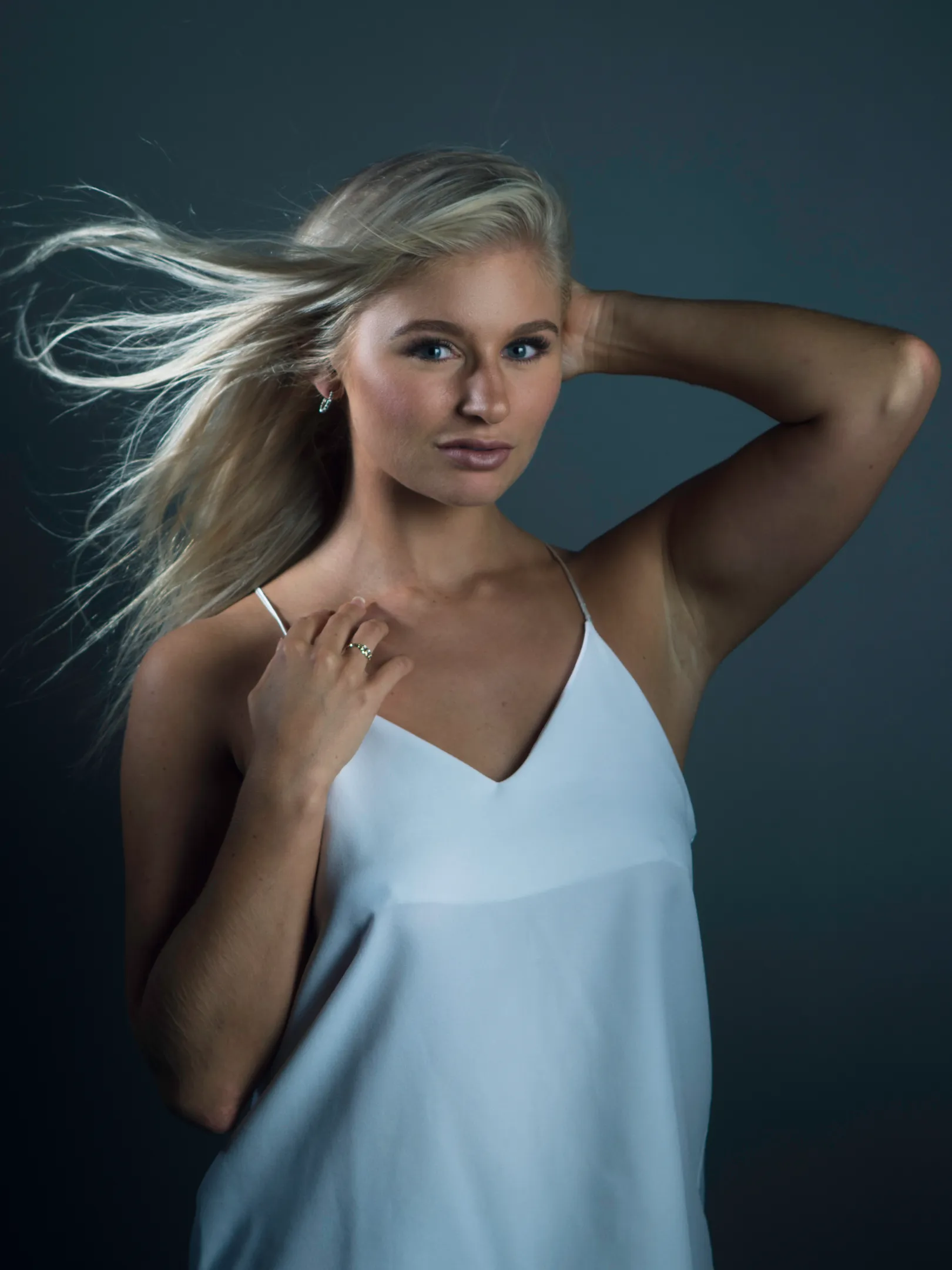Emily with long blonde hair wearing a white sleeveless top, posing with one hand on her head.