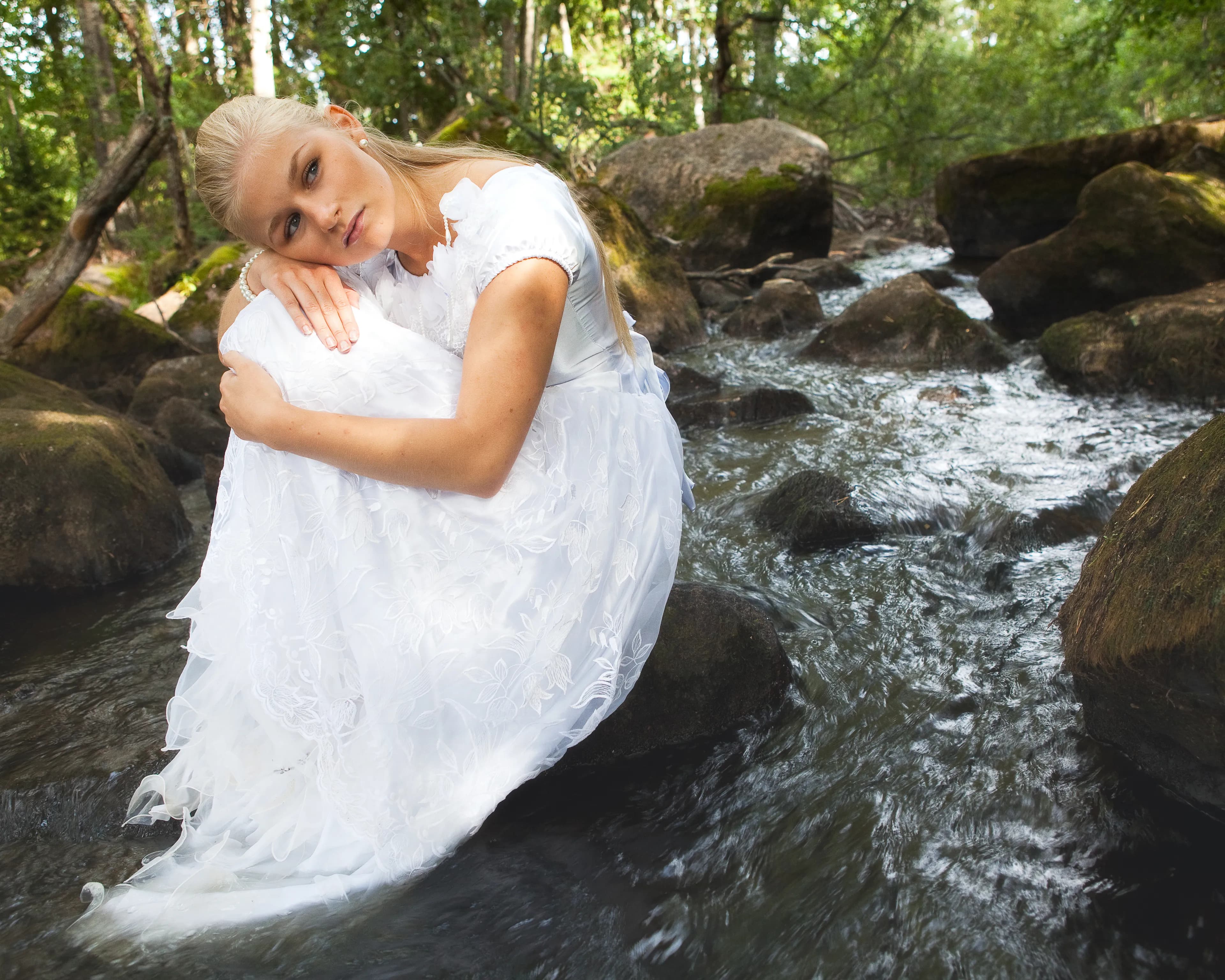 Emily in a white dress sitting by a forest stream.