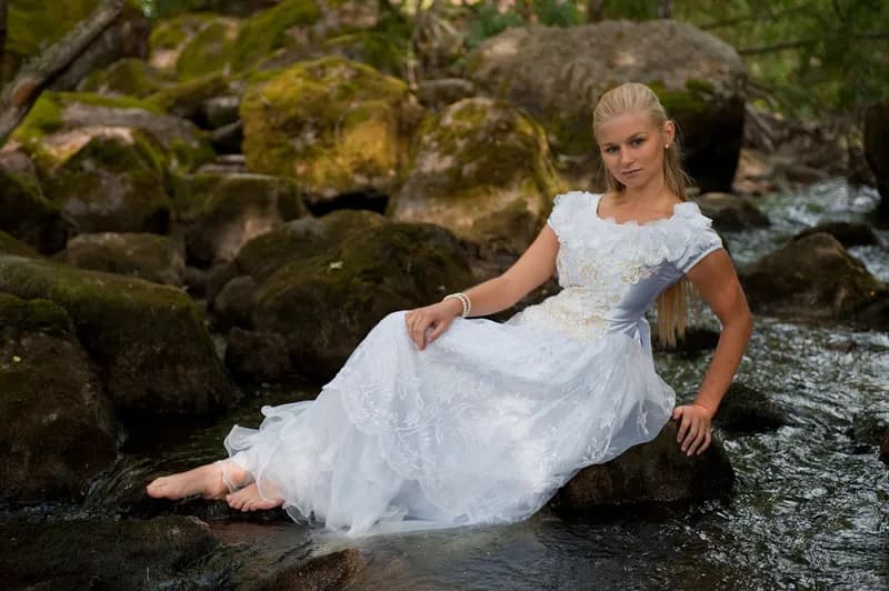 Emily in a white dress sitting on rocks by a stream in a forest.