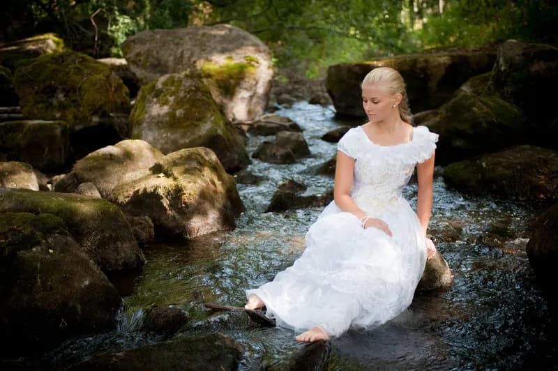 Emily in a white dress sitting on rocks by a stream in a forest.