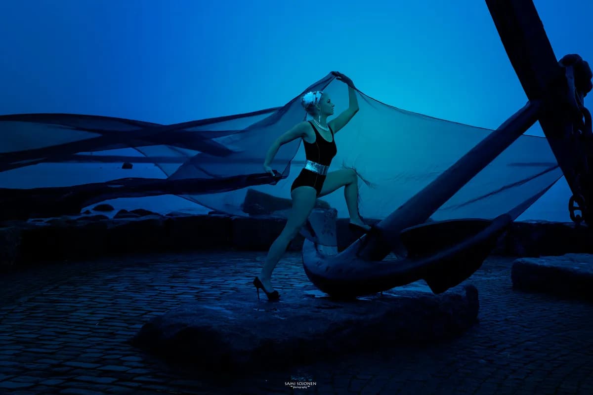 Emily posing dramatically in a black swimsuit with a flowing cape near a large anchor.