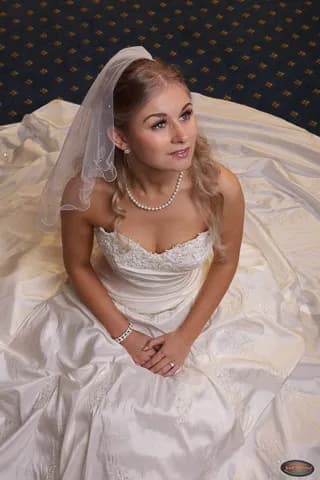 Emily in a wedding dress with a veil, sitting on a white satin fabric.
