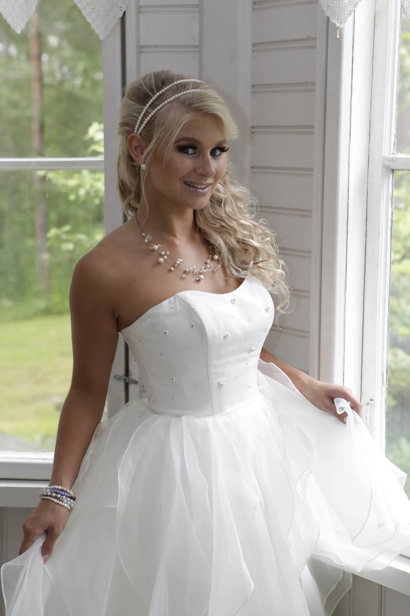 Emily in a white strapless wedding dress with a pearl necklace, standing by a window.