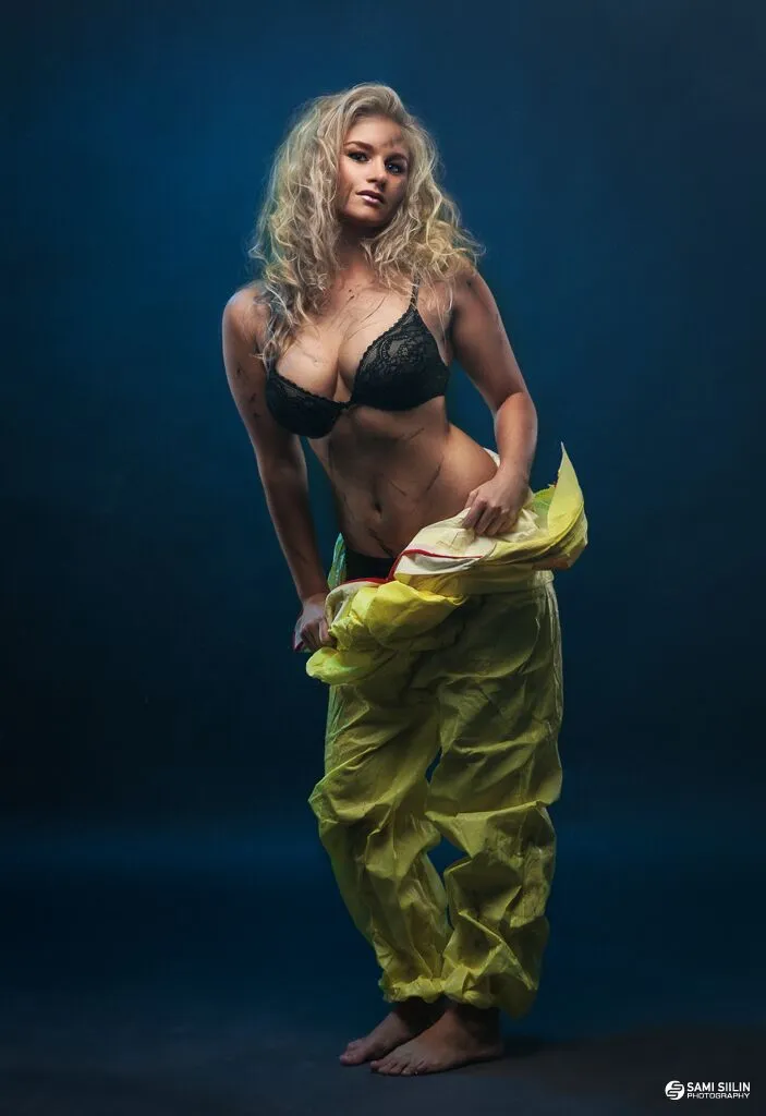 Emily posing in a black bra and yellow pants against a dark background.