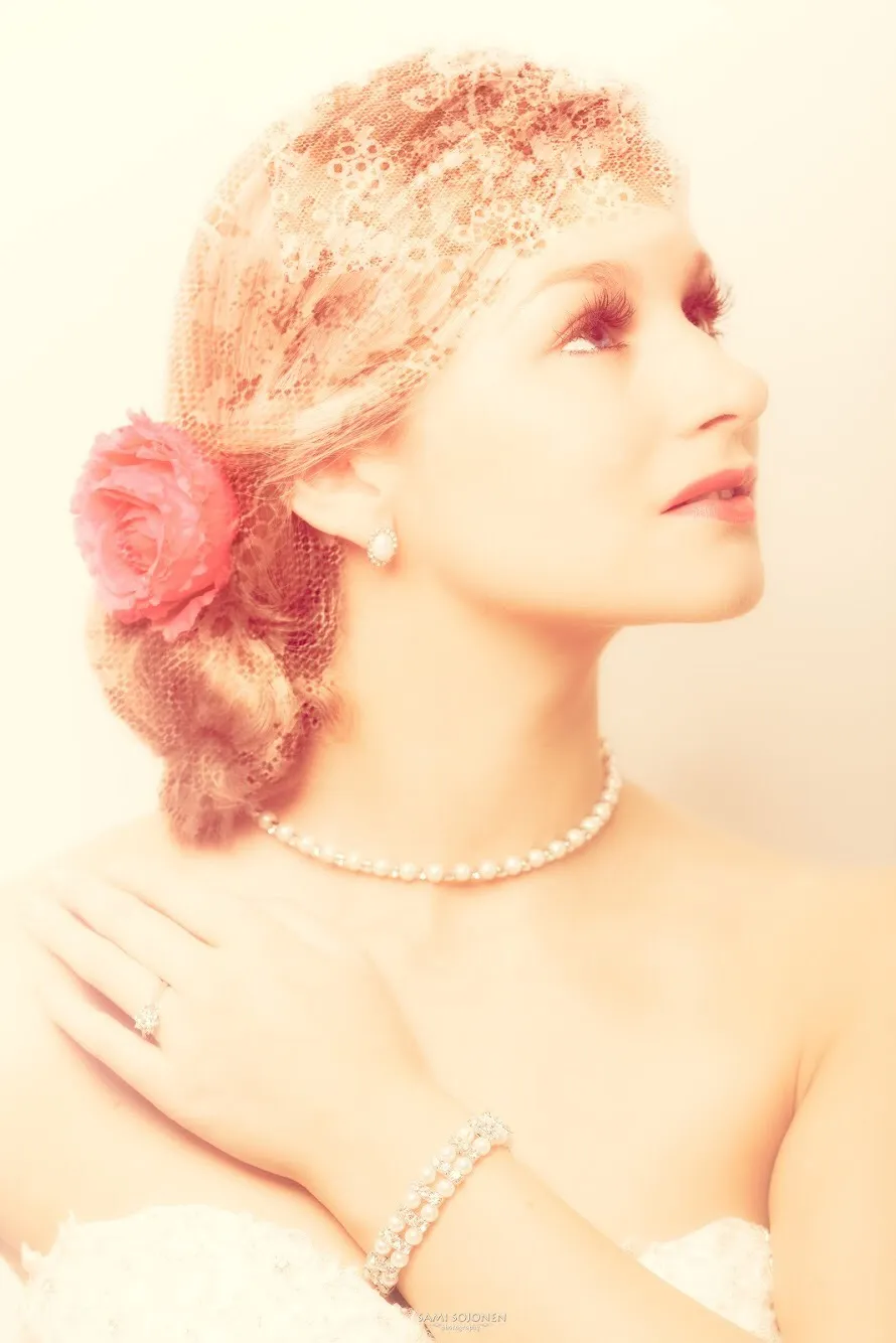 Emily in a vintage-style portrait with a lace veil and a red flower in her hair, wearing pearl jewelry.