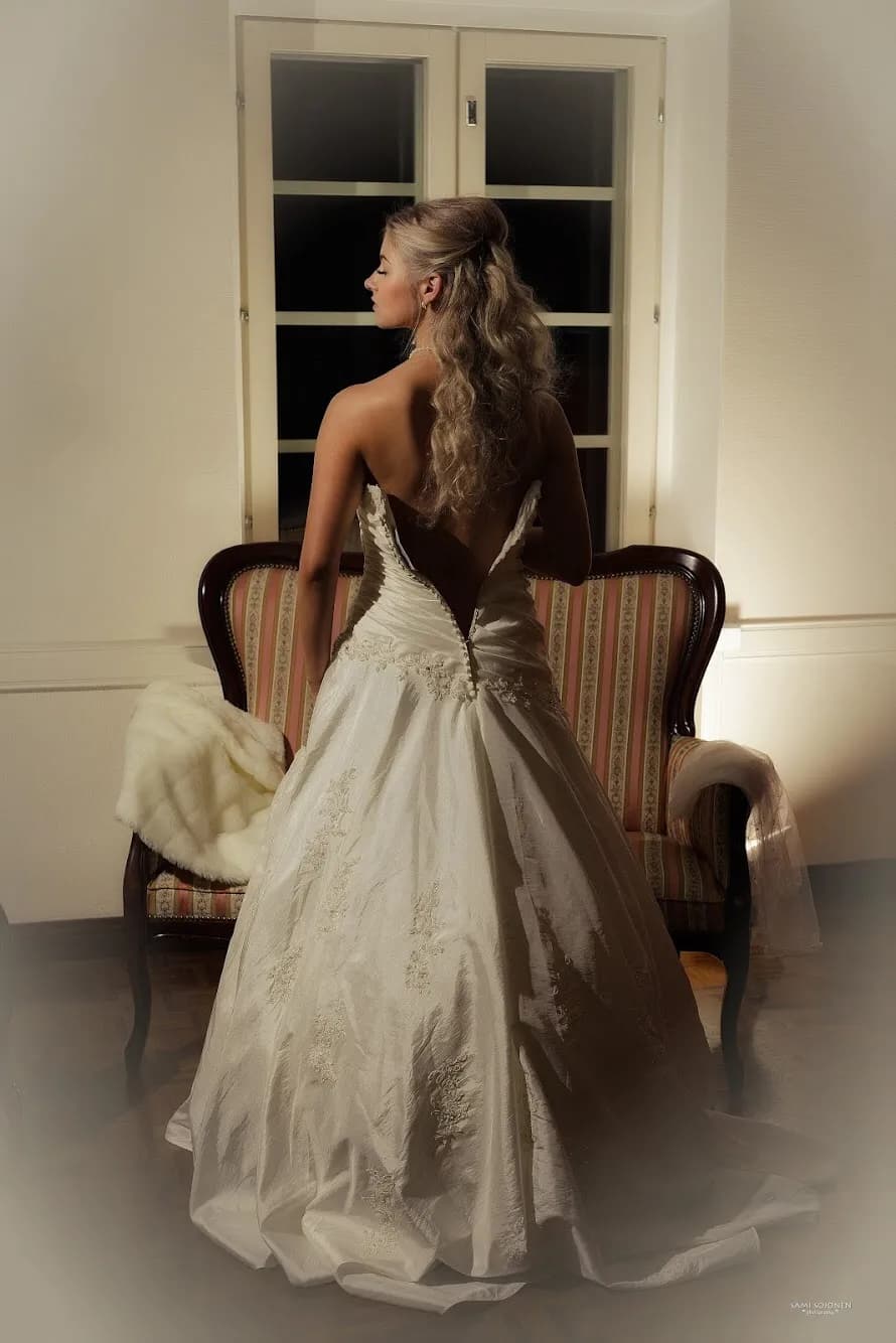 Emily in a white wedding gown with an open back, standing in front of a vintage chair.