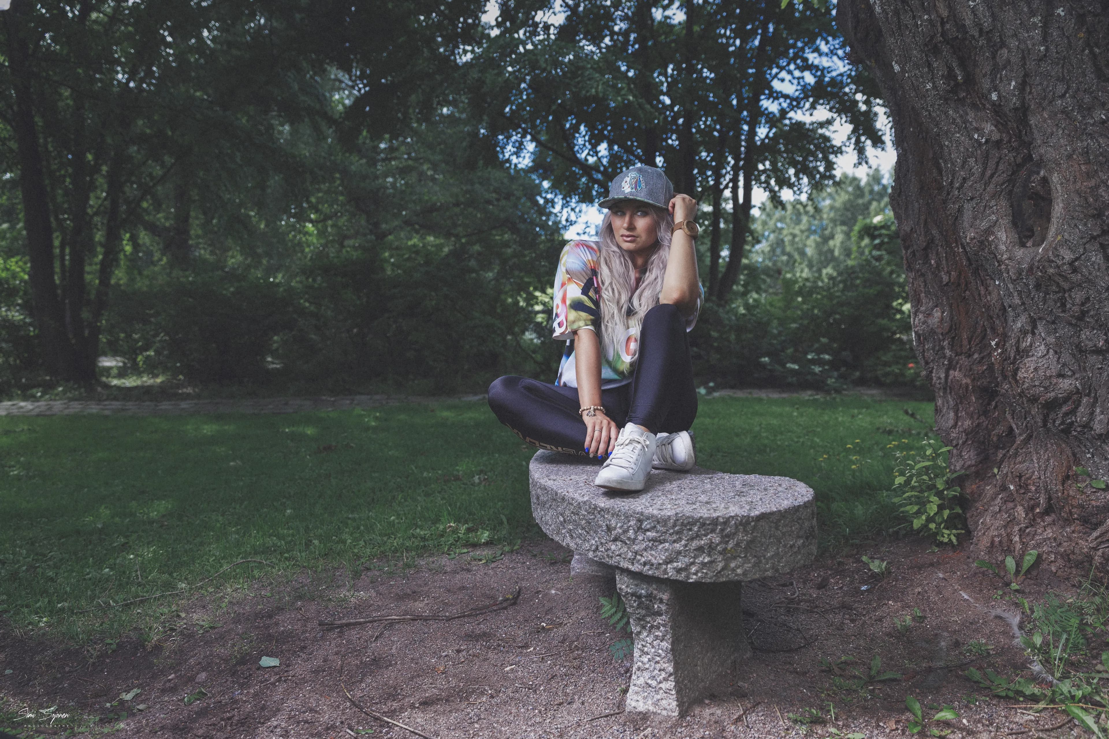 Emily sitting on a stone bench in a park, wearing a colorful shirt, black pants, white sneakers, and a cap.