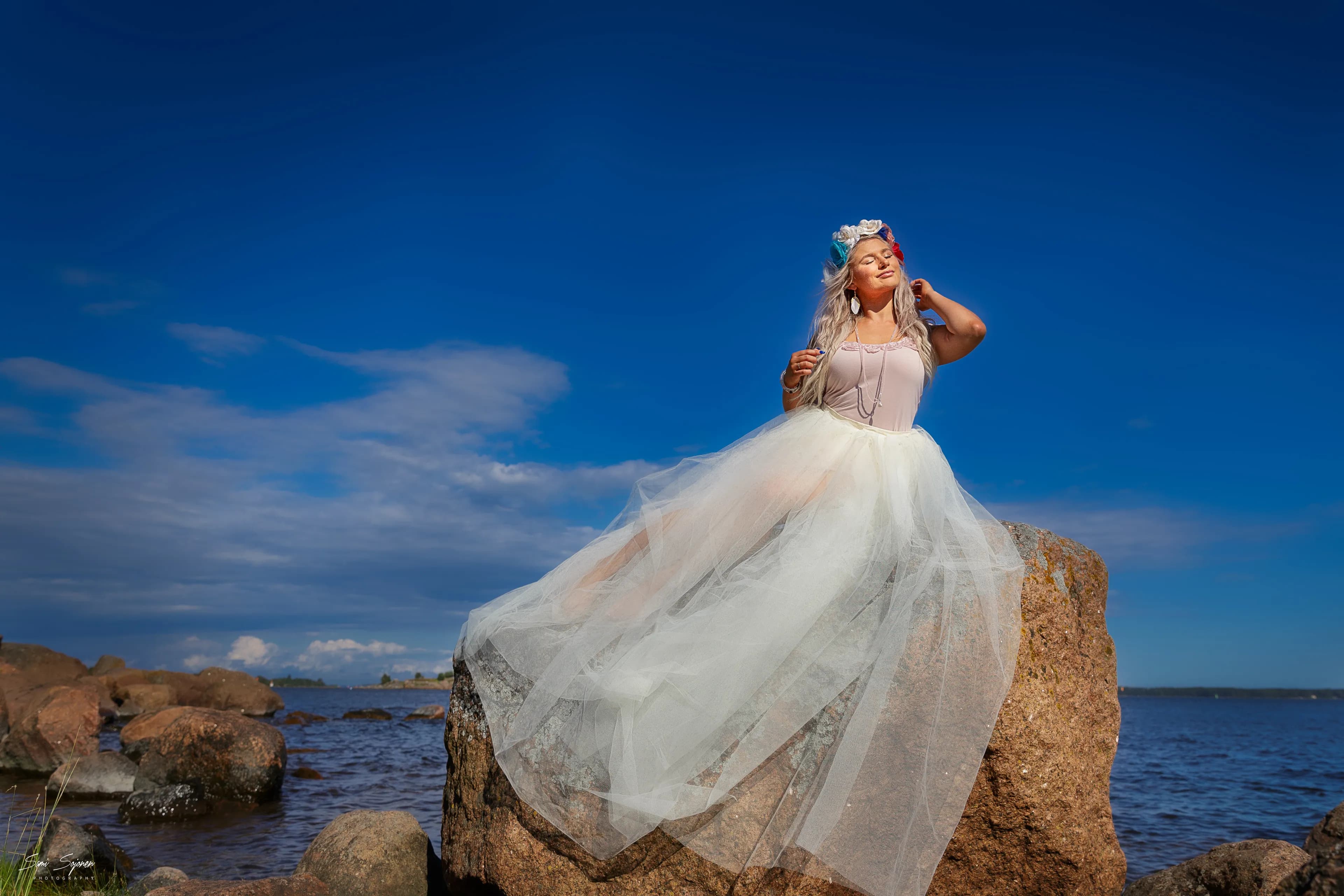 Emily in a flowing white dress poses on a large rock by the sea under a clear blue sky.