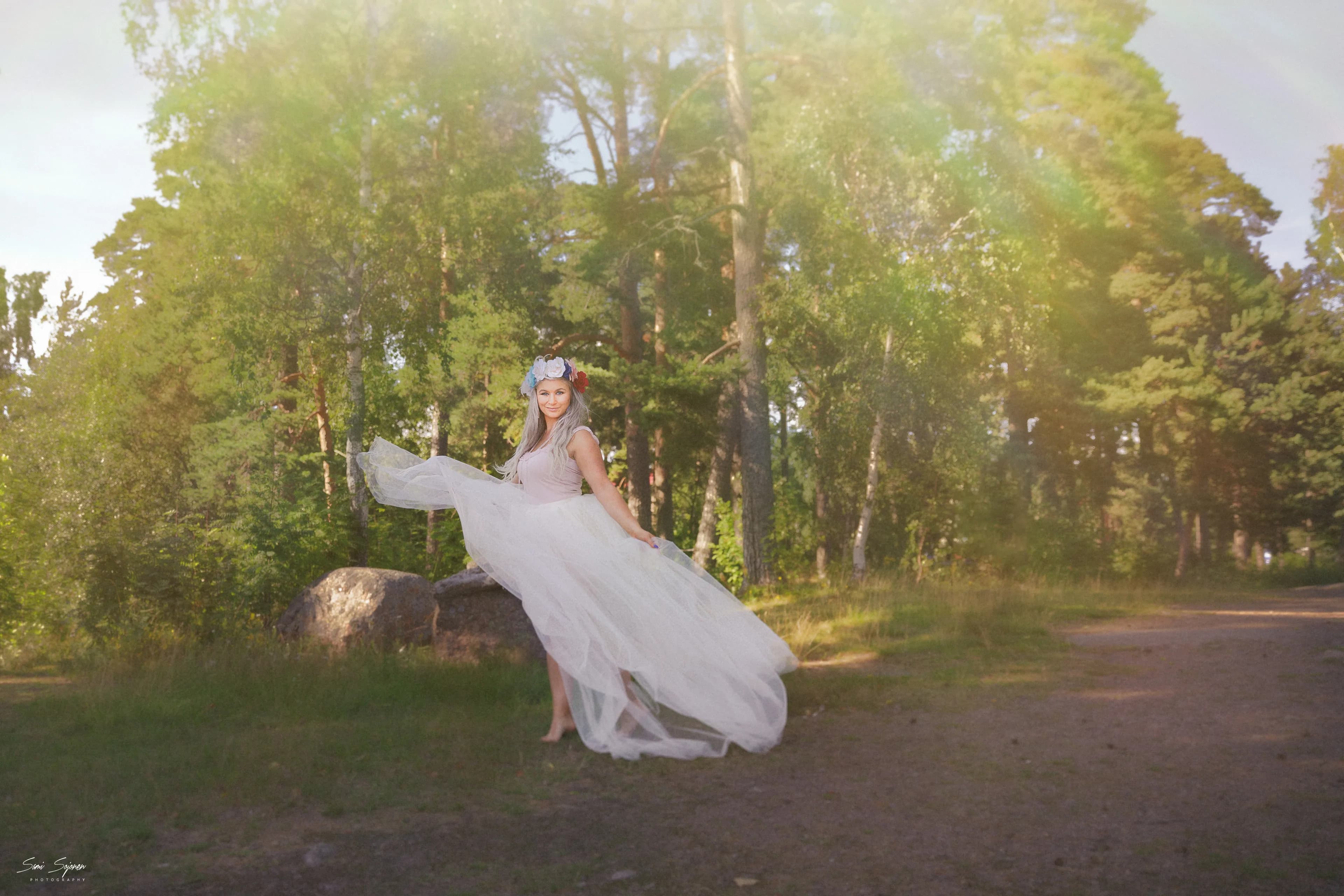 Emily in a flowing white dress standing in a forest with sunlight filtering through the trees.