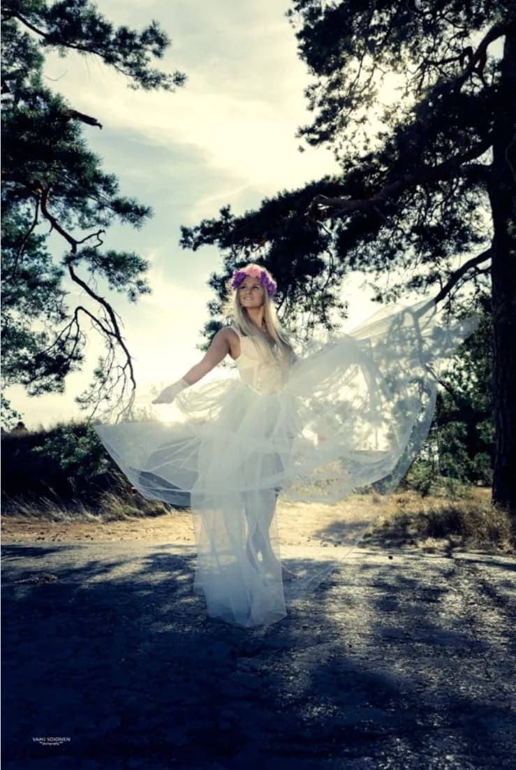 Emily wearing a flowing white dress and a flower crown, standing outdoors with trees and a bright sky.