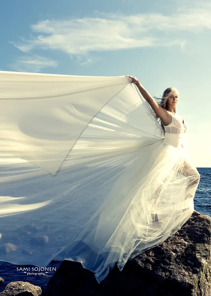 Emily in a flowing white dress standing on rocks by the sea.