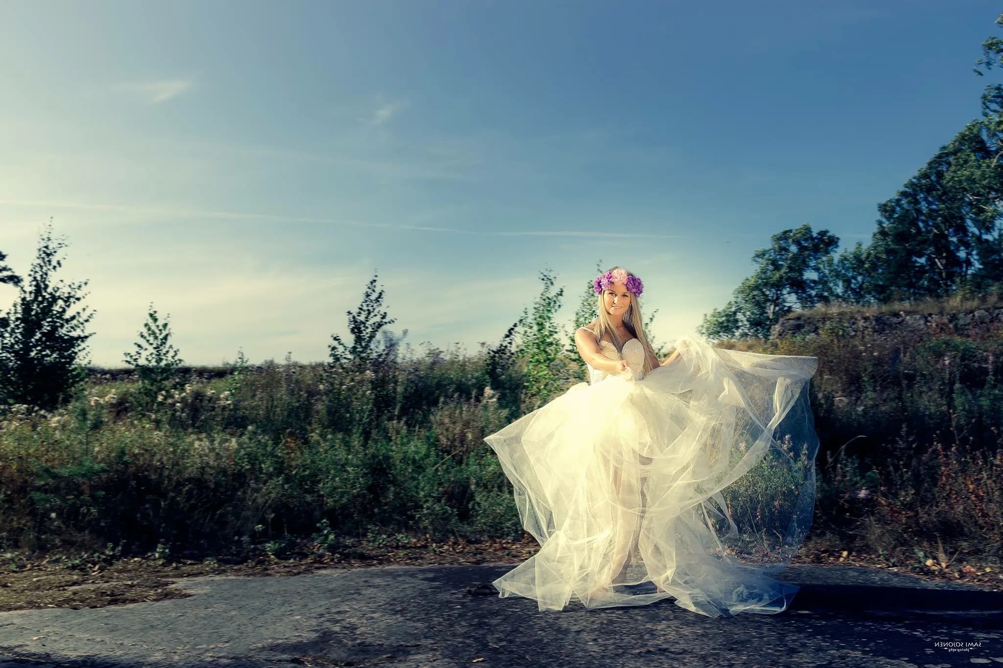 Emily in a flowing white dress with a floral headpiece, standing outdoors on a sunny day.
