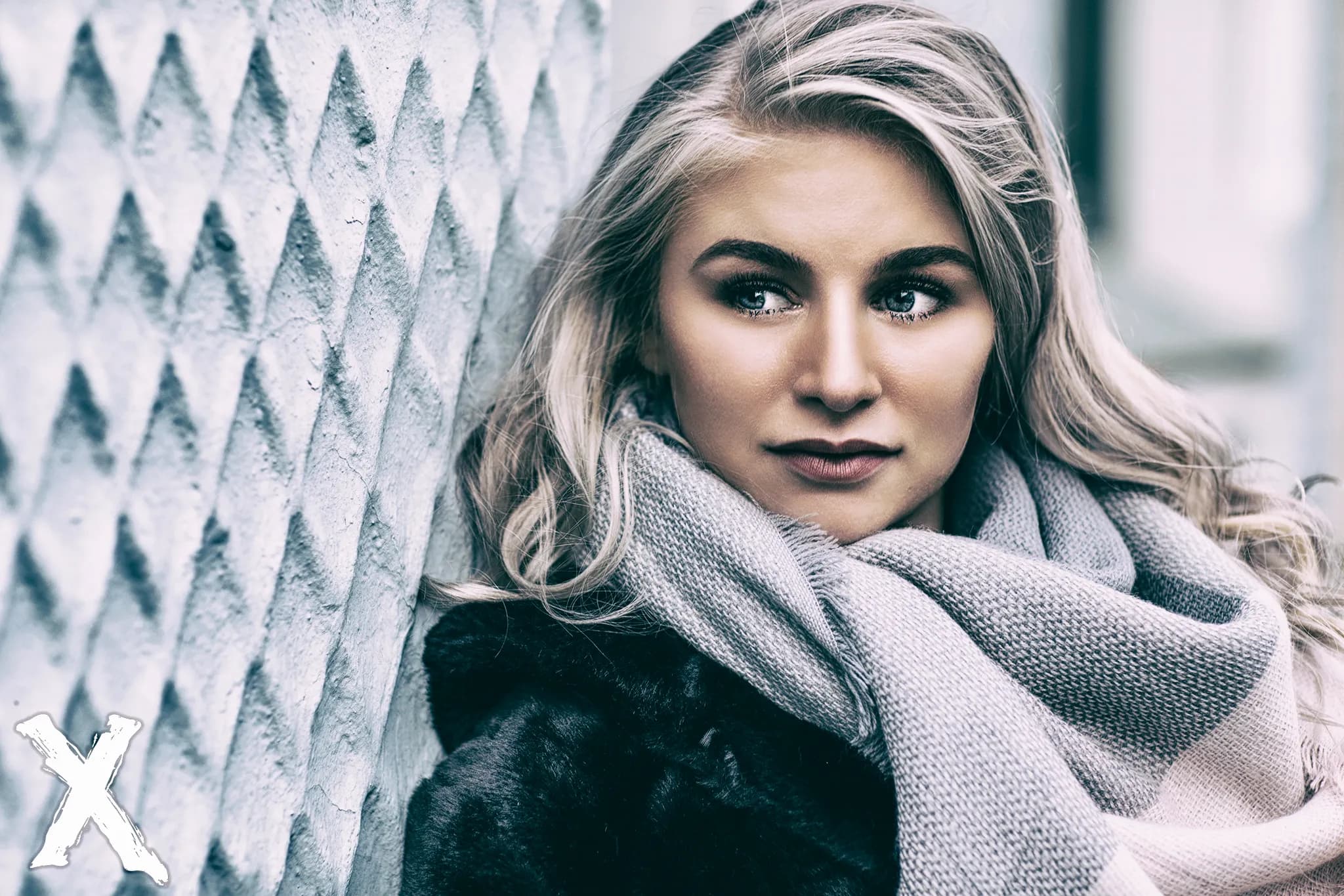 Emily with long blonde hair, wearing a scarf and coat, leaning against a textured wall.