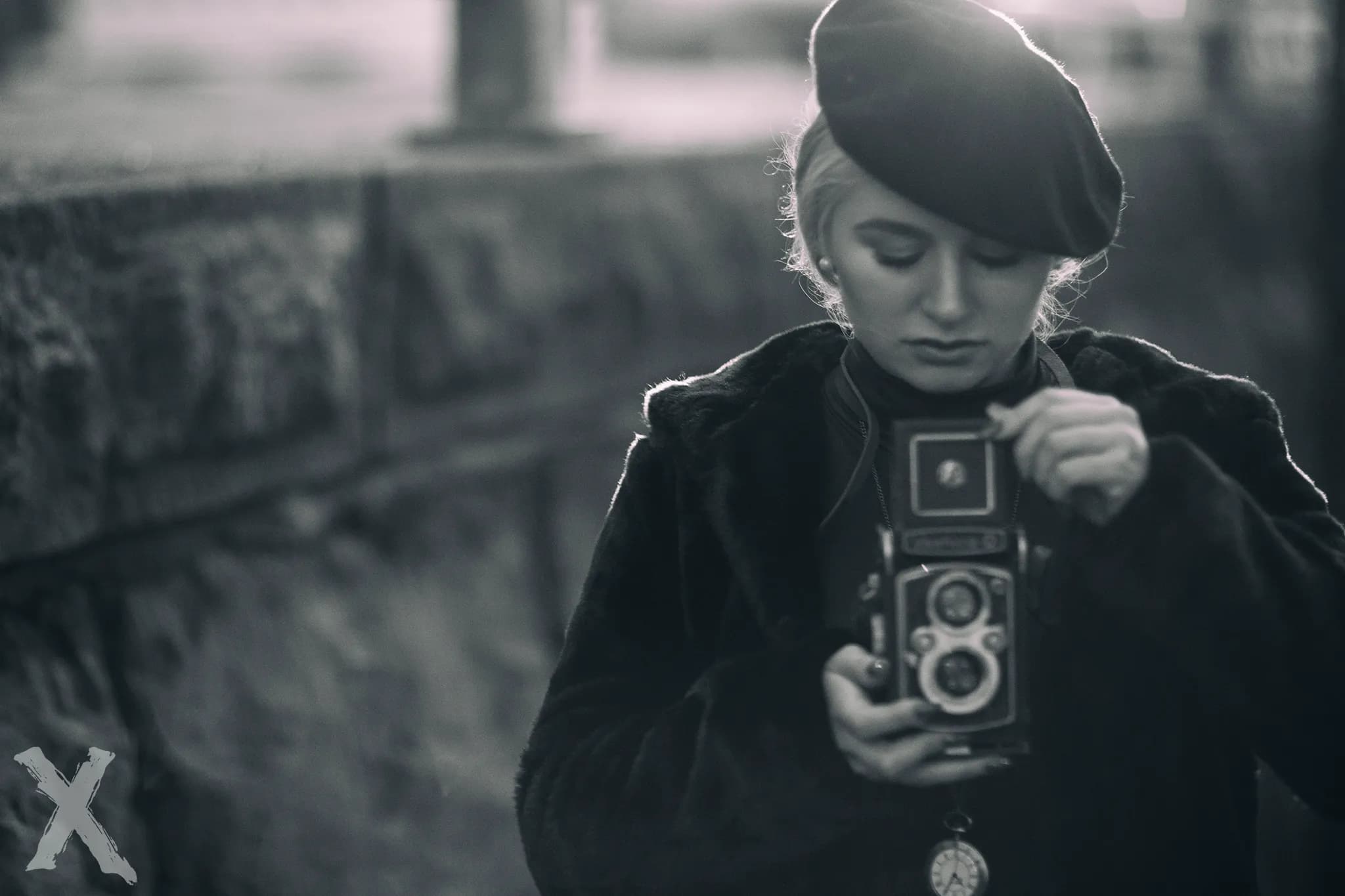 Emily, wearing a beret and coat, holding a vintage camera in a black and white setting.