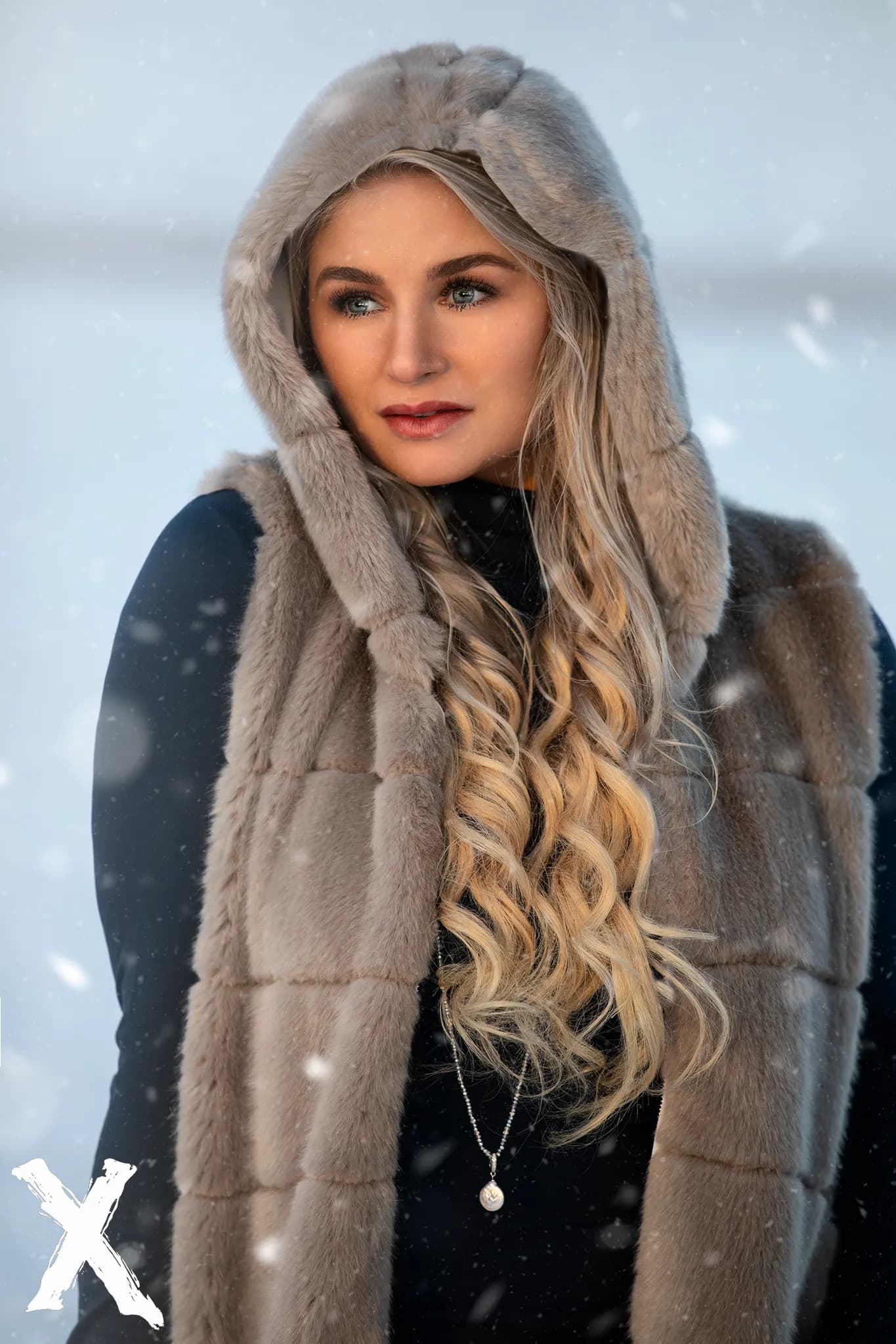 Emily wearing a fur hooded vest with long, wavy blonde hair, standing in a snowy setting.