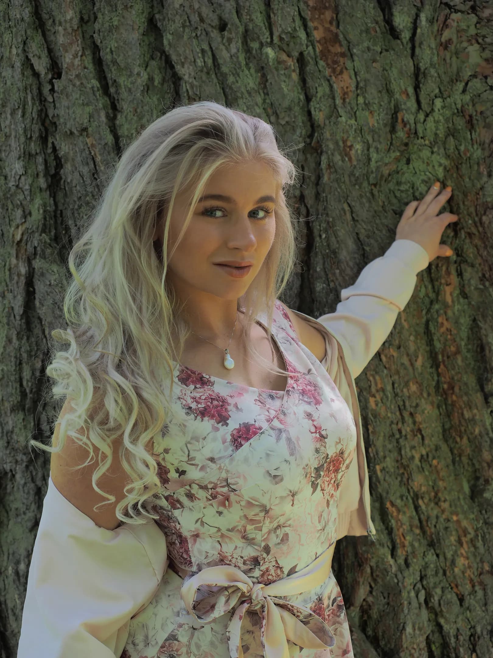 Emily with long blonde hair, wearing a floral dress, standing against a tree.