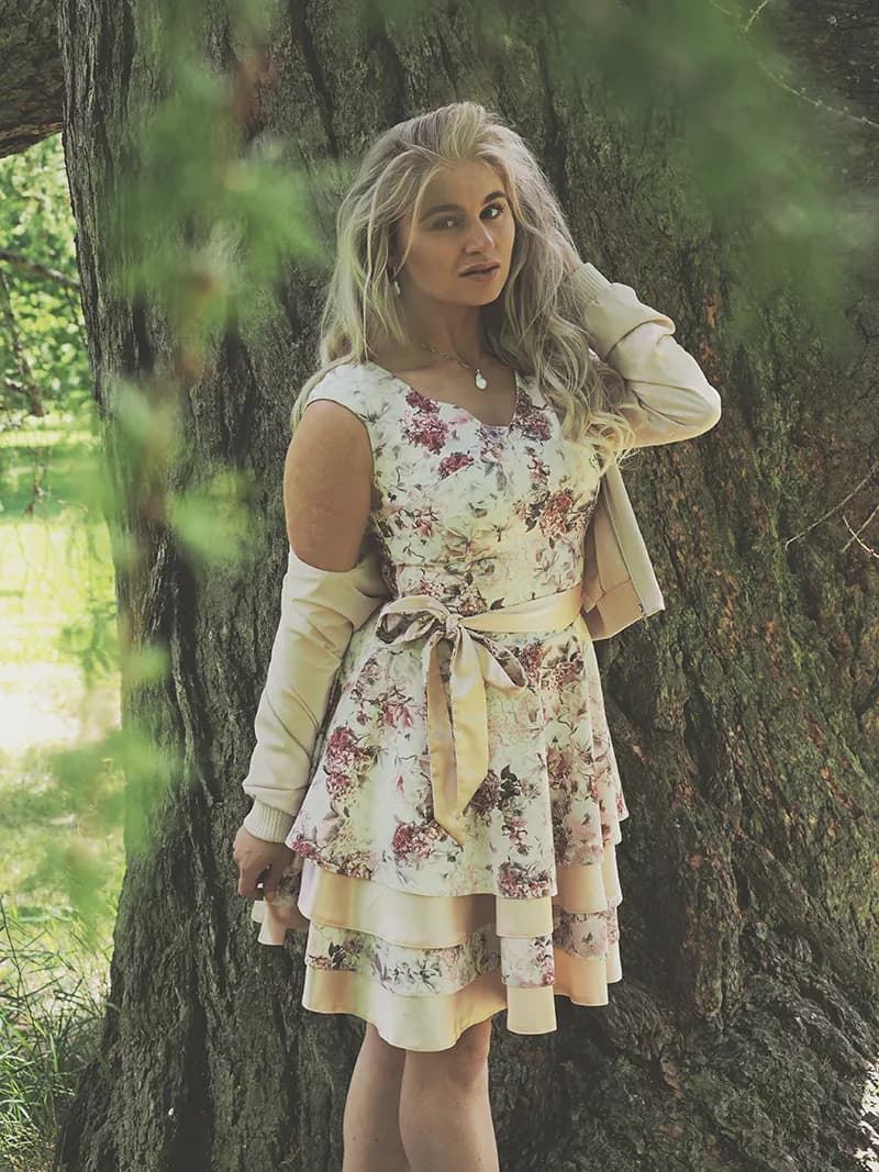 Emily standing by a tree wearing a floral dress and a light jacket.