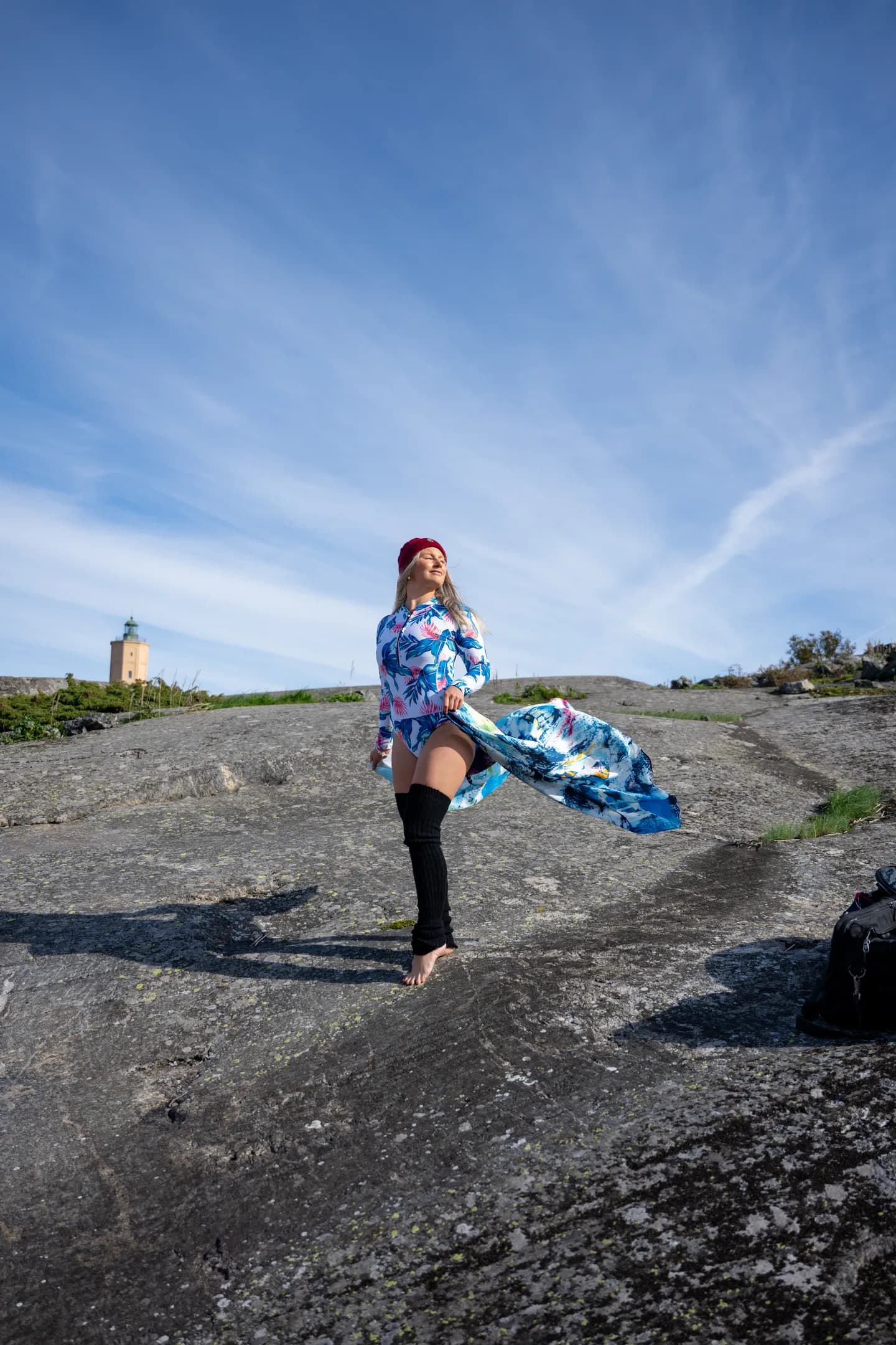 Emily standing on a rocky surface wearing a colorful dress and a red hat, with a flowing scarf in the wind.