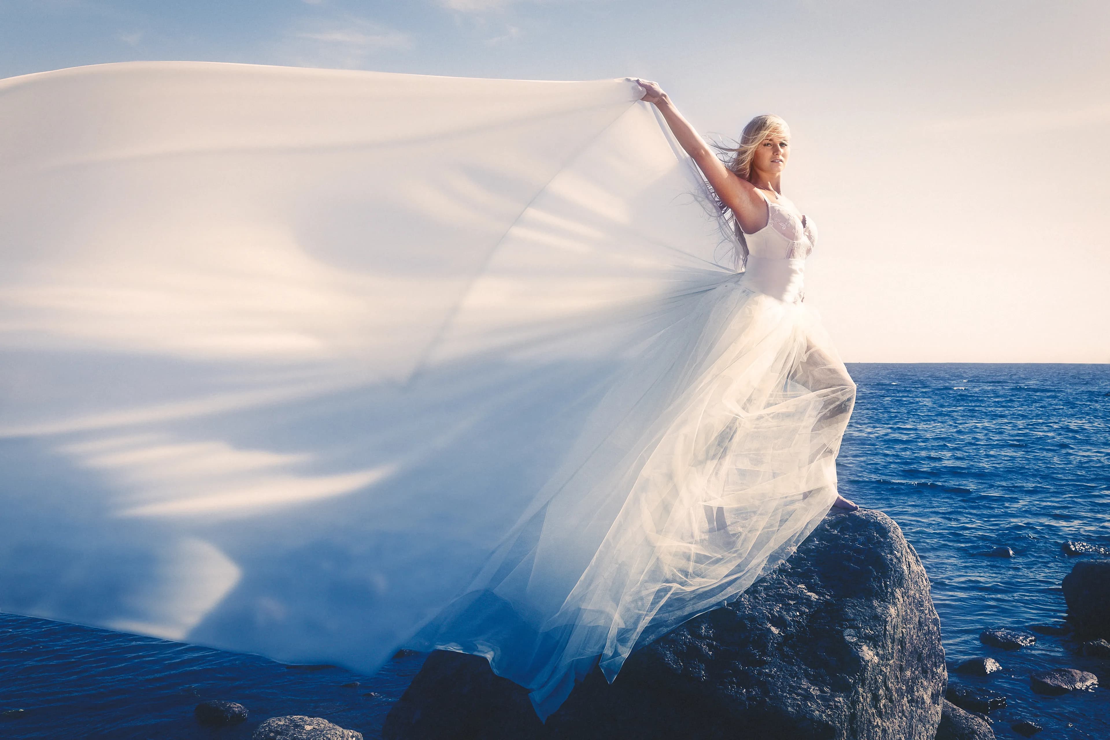 Emily in a flowing white dress standing on rocks by the ocean.