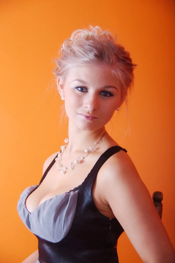 Emily posing against an orange background, wearing a black and gray dress with a pearl necklace.