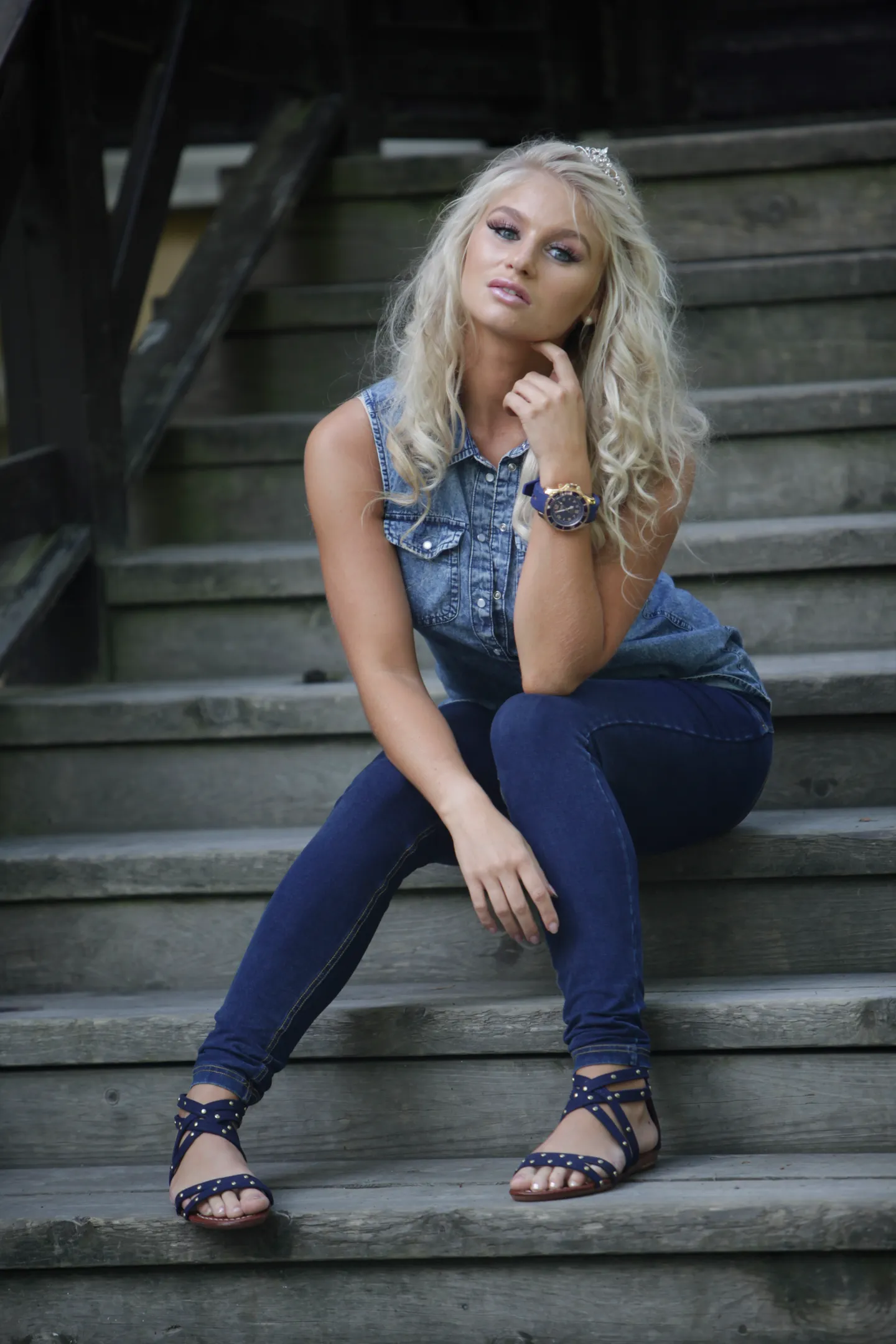Emily sitting on wooden stairs, wearing a denim outfit and sandals.