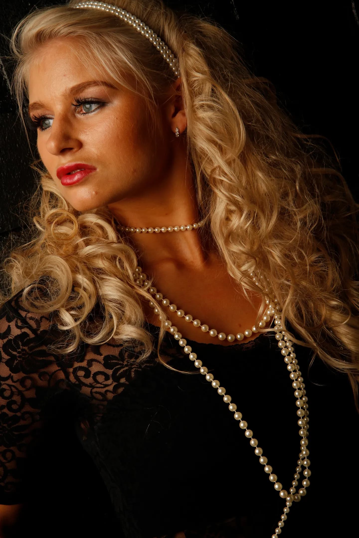 Emily with long curly blonde hair, wearing pearl jewelry and a black lace top.