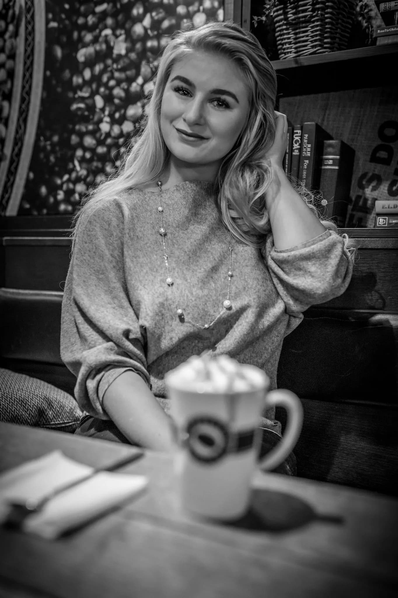 Emily sitting in a cozy cafe with a warm smile, wearing a sweater and a necklace, with a cup of coffee in the foreground.